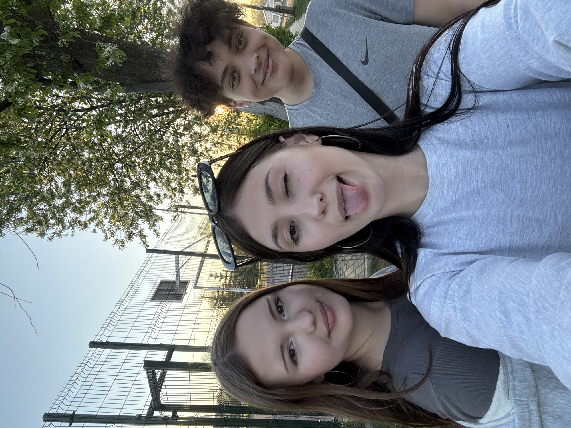 Three people are posing for a photo outdoors near a fence, trees, and possibly a building. The person at the bottom of the image has light brown hair and is smiling. The person in the middle has dark hair, is winking, and sticking their tongue out. The person at the top has curly hair and a gray Nike shirt. A black strap is visible across the upper person's chest. The scene appears to be in the city of Фоки, Russia, although specific location details are not evident from the image.