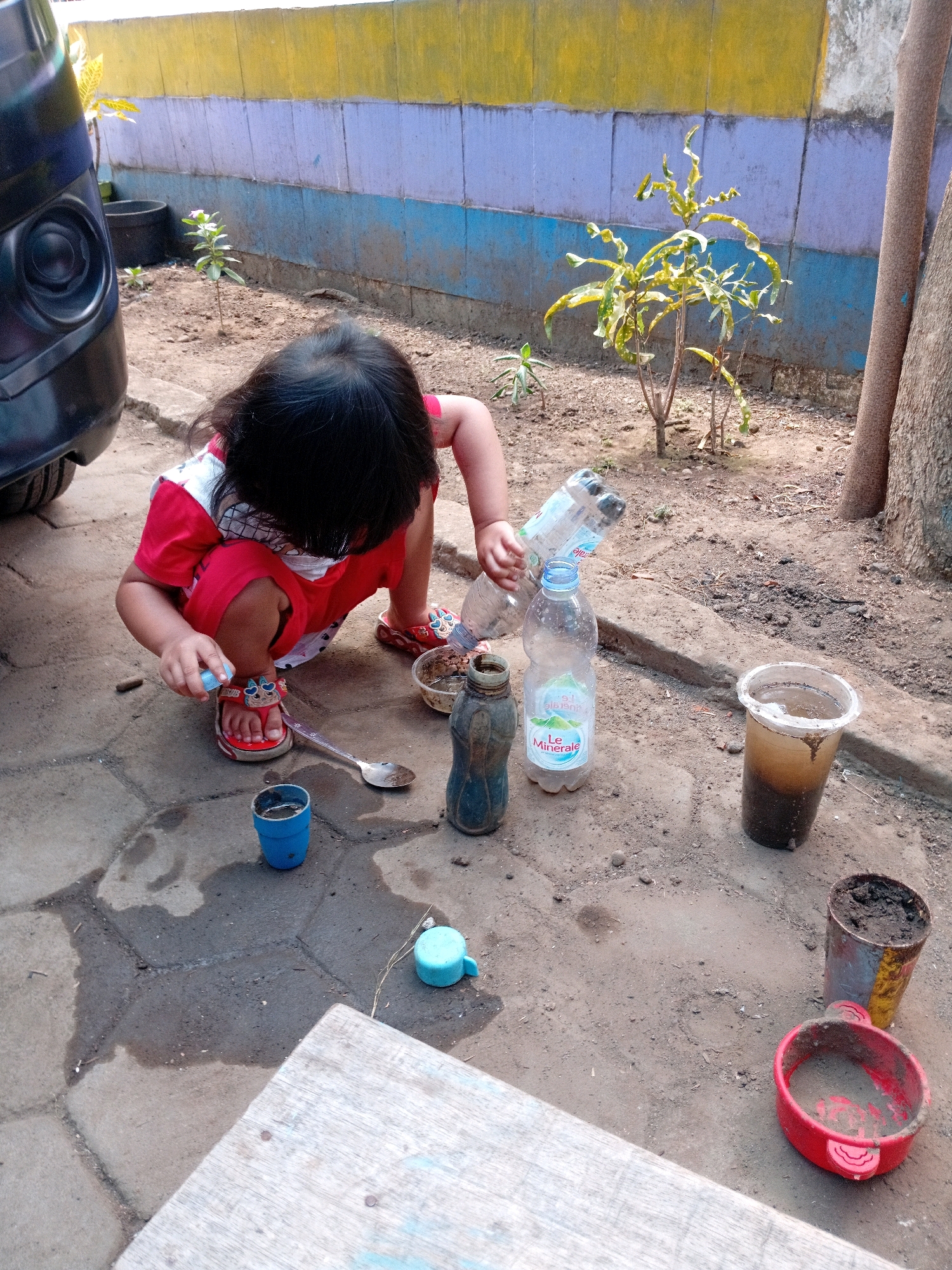 An Indonesian child with black hair and wearing a red shirt and red sandals is crouching on a paved area outside near a wall. The wall has horizontal stripes of yellow, purple, and blue. The child is reaching for a small blue cup with their left hand, while their right hand is holding a small blue bottle. In front of the child are various objects, including a silver spoon, two plastic bottles (one with a white label that reads "Le Minerale"), a dark-colored bottle, and a clear plastic cup containing dark liquid and sediment. To the right of these objects, there are two cylindrical containers, one with soil and a yellow stripe and another red bowl with soil. The ground is a mixture of dirt and paving stones, with some puddles of water visible. A portion of a black vehicle is visible on the left side of the image. A tree trunk is visible on the right.