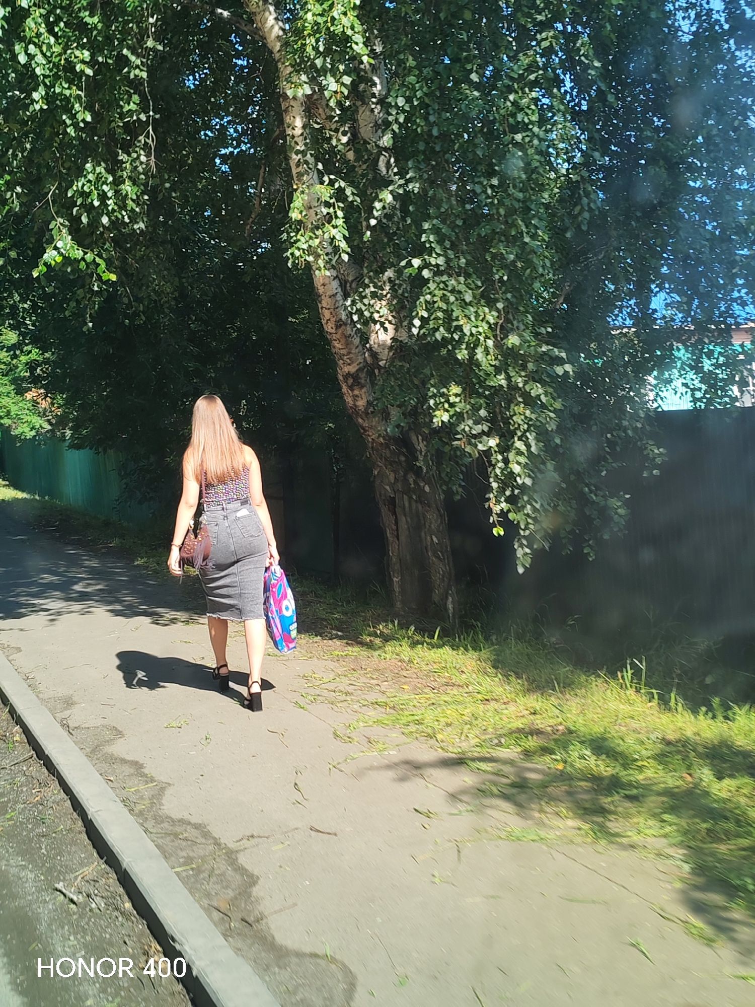 A woman with long, blonde hair, seen from behind, walks along a paved path next to a grass verge. She is wearing a patterned tank top and a gray denim skirt. In her right hand, she carries a colorful tote bag. Her left hand is clasped around a brown purse with a fringe detail. She is wearing black high-heeled sandals. The path is bordered by a concrete curb on the left. A lush green fence runs along the right side of the path, with trees and foliage visible beyond it. The scene appears to be set on a sunny day, with dappled sunlight and shadows cast on the path and grass. The lower left corner of the image displays the text "HONOR 400". The location is identified as Bratsk, Russia.
