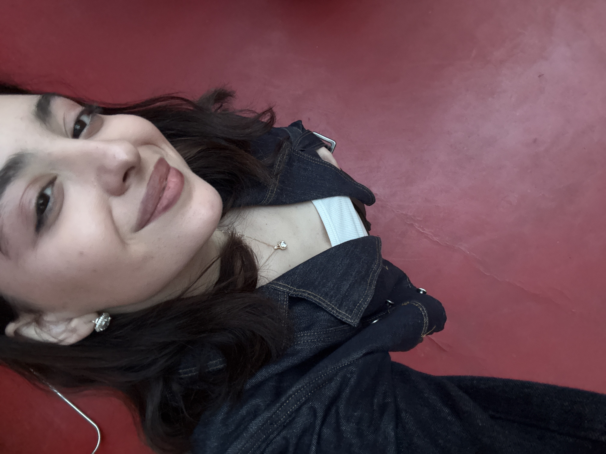 A woman is pictured from a high angle against a red background. She wears a dark denim jacket over a white top, with a delicate necklace and earrings visible. The location is Yerevan, Armenia, based on metadata.