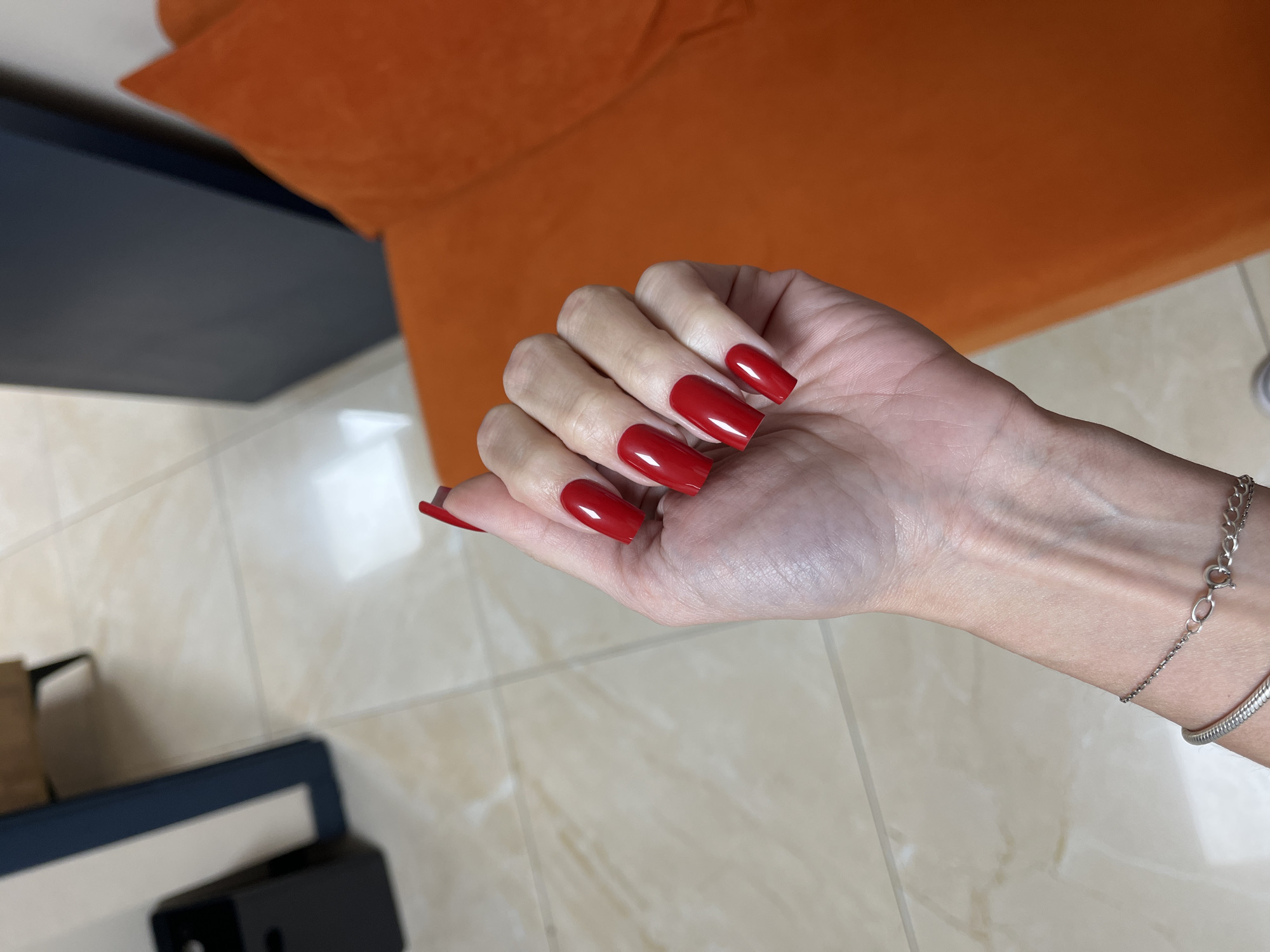 The image shows a person's hand with long, red, square-shaped fingernails. The hand is resting on a tiled floor with a marble-like pattern. In the background, a portion of an orange couch is visible, along with a dark blue surface. A silver bracelet with a clasp is worn on the wrist. The overall setting appears to be indoors, possibly a living room or a lounge area, in Nizhny Novgorod, Russia. The lighting suggests daytime. There is no visible text in the image. The focus is on the hand and its adornments.