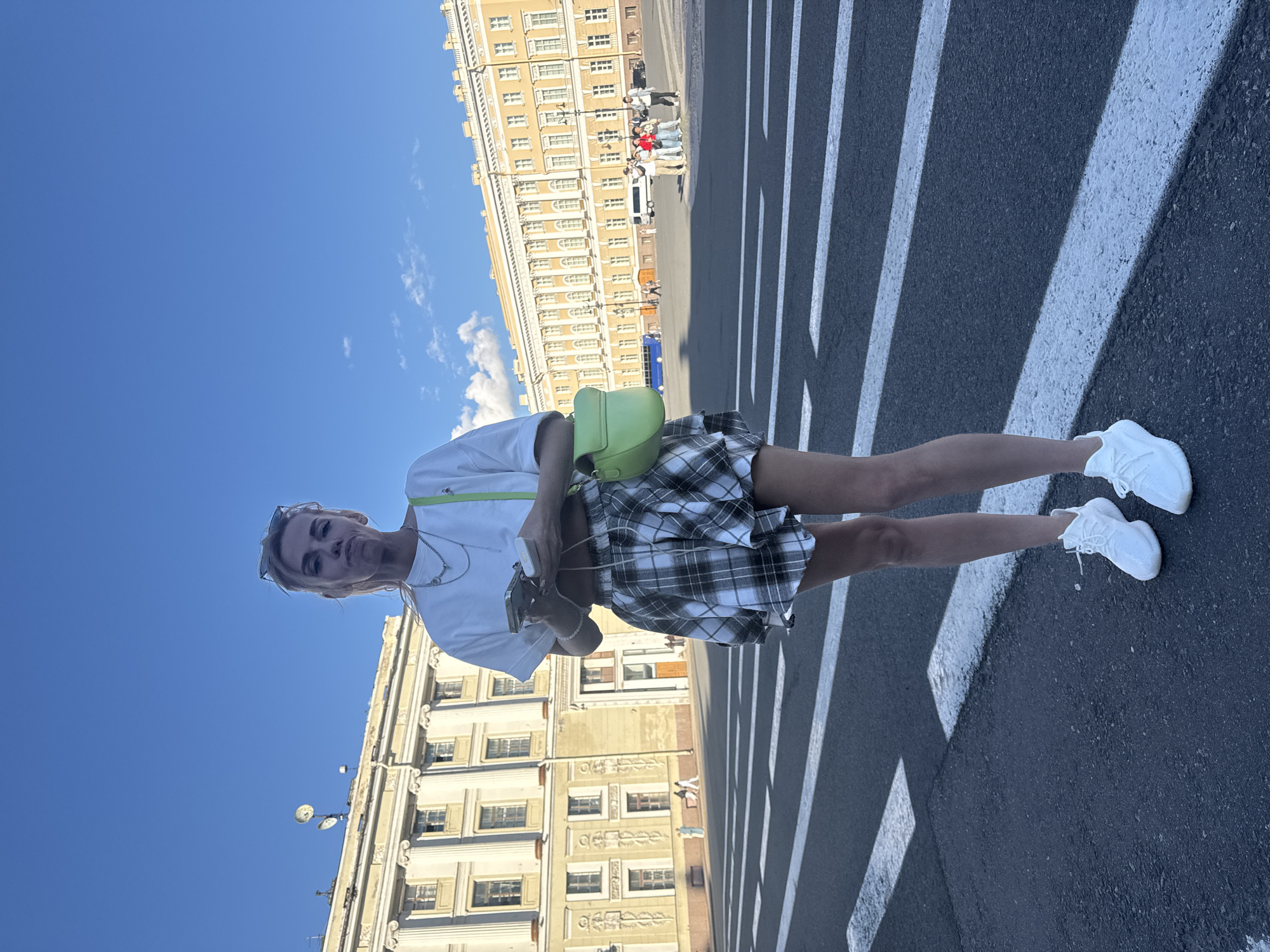 The media file shows a young woman standing on a pedestrian crossing in Saint Petersburg, Russia. She is wearing a white t-shirt, a plaid skirt, and white sneakers. A bright green fanny pack is slung across her chest. She is holding a smartphone in her right hand and looking at the camera with a neutral expression.
In the background, a large, ornate building with many windows and classical architectural details lines the street. The sky is a clear, vibrant blue with a few wispy clouds. The sun is high in the sky, casting distinct shadows from the pedestrian crossing lines onto the asphalt, suggesting it is daytime, likely mid-day. Other people are visible in the distance, some walking and others gathered near the building, implying a public and likely tourist area. The overall scene conveys a sense of urban exploration on a bright, sunny day.