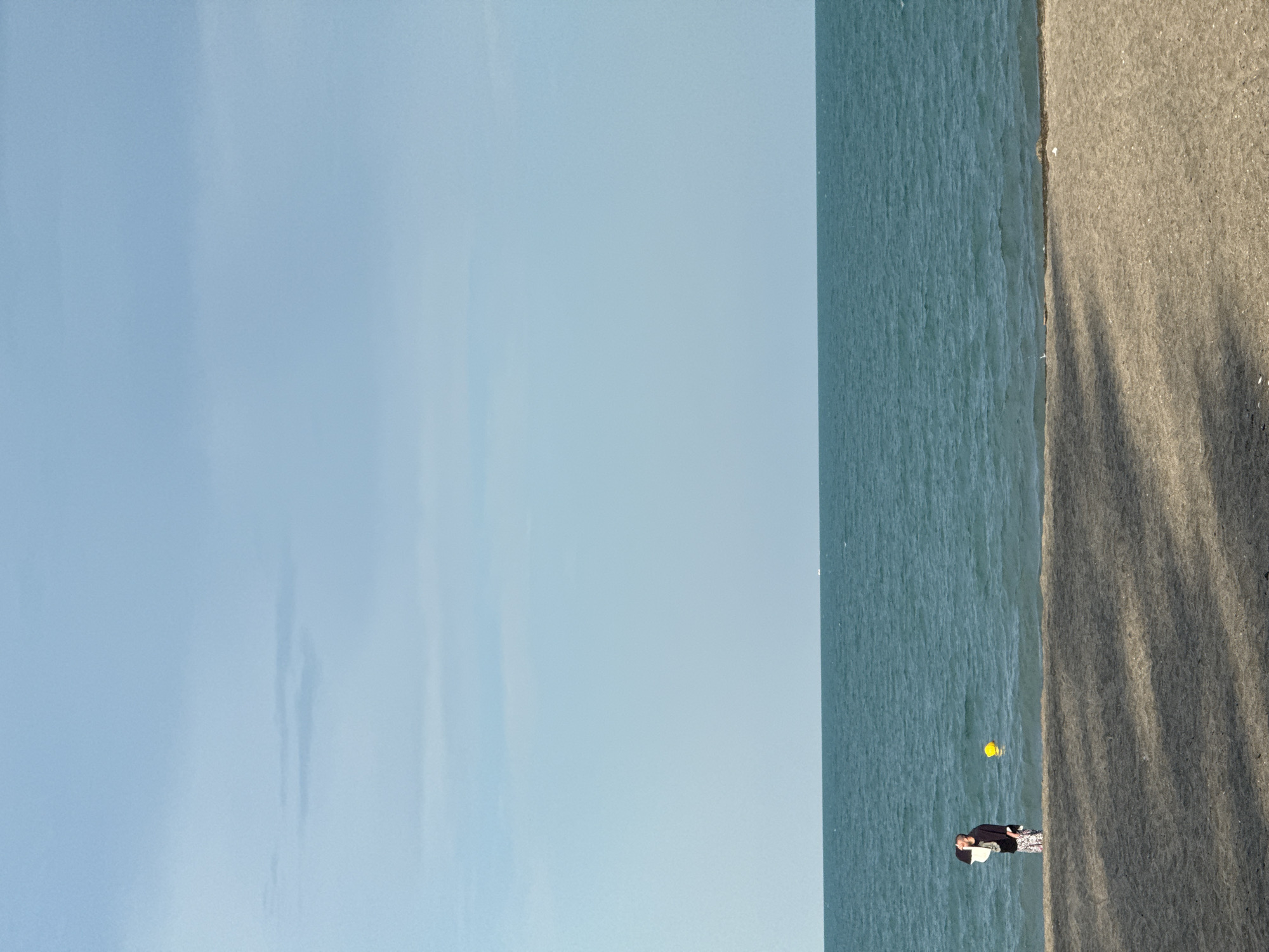 The image shows a beach scene. On the right side of the frame is a sandy beach, casting long shadows from an unseen light source, suggesting it is late afternoon or early morning. The beach meets the ocean, which occupies the middle section of the image. The water is a deep blue-green and appears calm with gentle waves. In the water, a yellow buoy is visible. On the sandy beach, a person is lying down, facing away from the camera. They are wearing dark clothing and patterned leggings. The person appears to be relaxed or sleeping. The left side of the image is dominated by a pale blue sky, with some faint cloud formations. There is no visible text in the image. The scene depicts a tranquil moment on the coast.