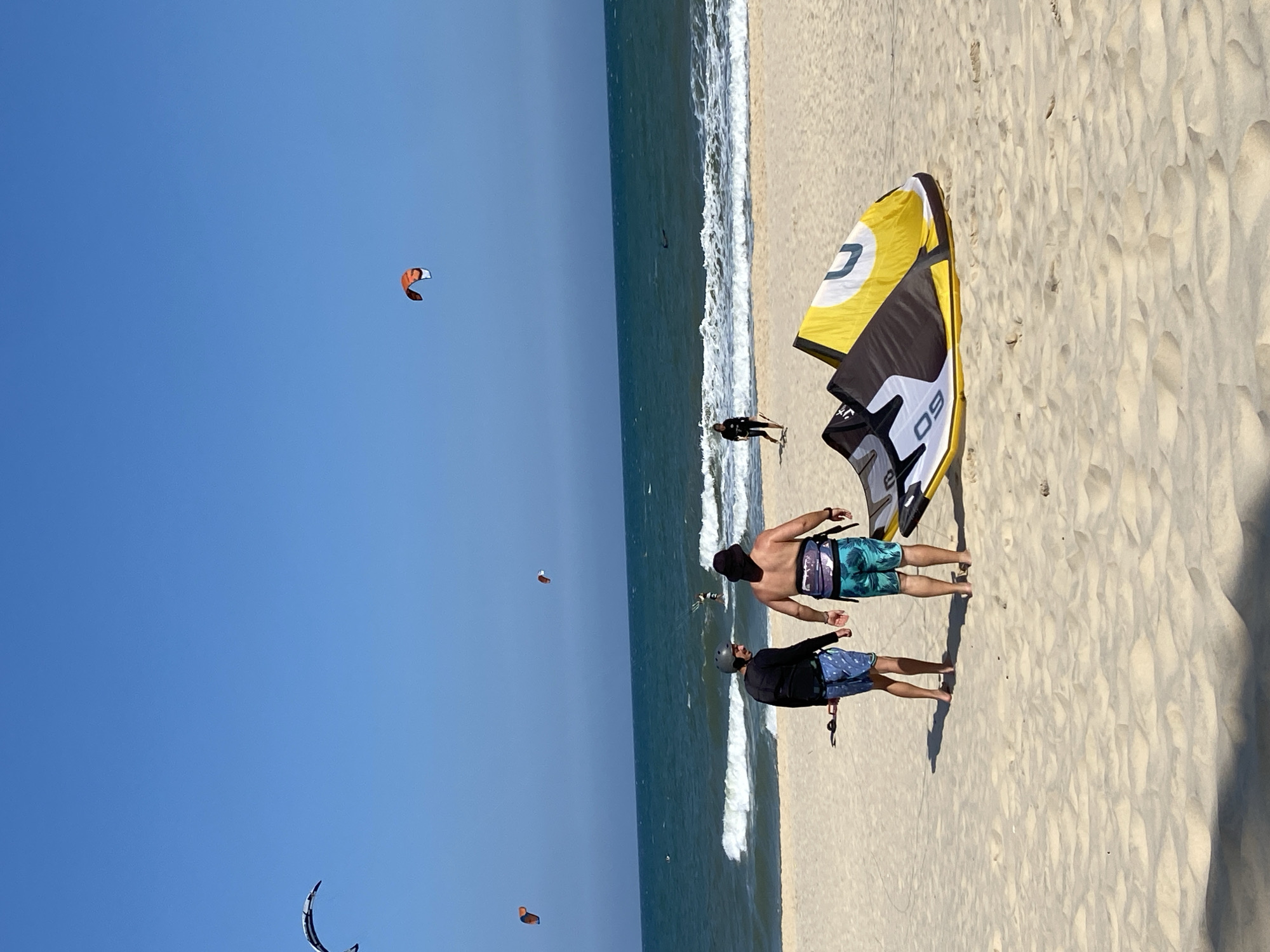 Under a clear blue sky at a beach in Phu Thuy Ward, Vietnam, a kitesurfing scene is depicted. The light-colored sandy beach meets the ocean, where white foamy waves break. In the foreground, two men stand on the sand, facing the water. The man on the left wears a dark long-sleeved shirt, a grey helmet, blue patterned swim shorts, and a dark harness, holding a control bar. The man on the right, shirtless, wears a dark bucket hat, blue-green patterned swim shorts, and a dark harness. Adjacent to them on the sand are two kitesurfing kites: one primarily yellow, white, and black with a "DU" design, and another black, white, and grey kite marked "09". Further down the beach to the right, a person in dark clothing walks towards the water. In the ocean, several individuals engage in kitesurfing, with one actively riding a board. Four kites are airborne: a prominent black and white kite in the upper left, an orange and white kite in the upper right, and two smaller orange and white kites positioned further out over the water.