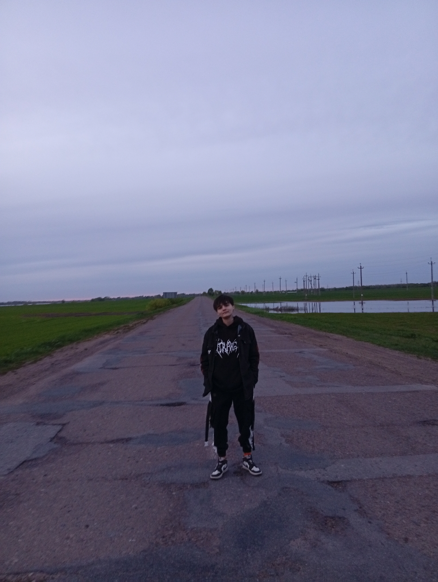 The photograph depicts a person standing on a paved road. The individual has short dark hair, and is wearing a black hooded sweatshirt with a graphic print on the front, black pants, a black jacket, and two-toned shoes. They are facing the camera with their hands in their pockets. The road appears to be cracked and uneven. On either side of the road, there is grass, and what appears to be standing water. In the background, telephone poles stretch into the distance beneath an overcast sky. The location is Azyakul', Russia.