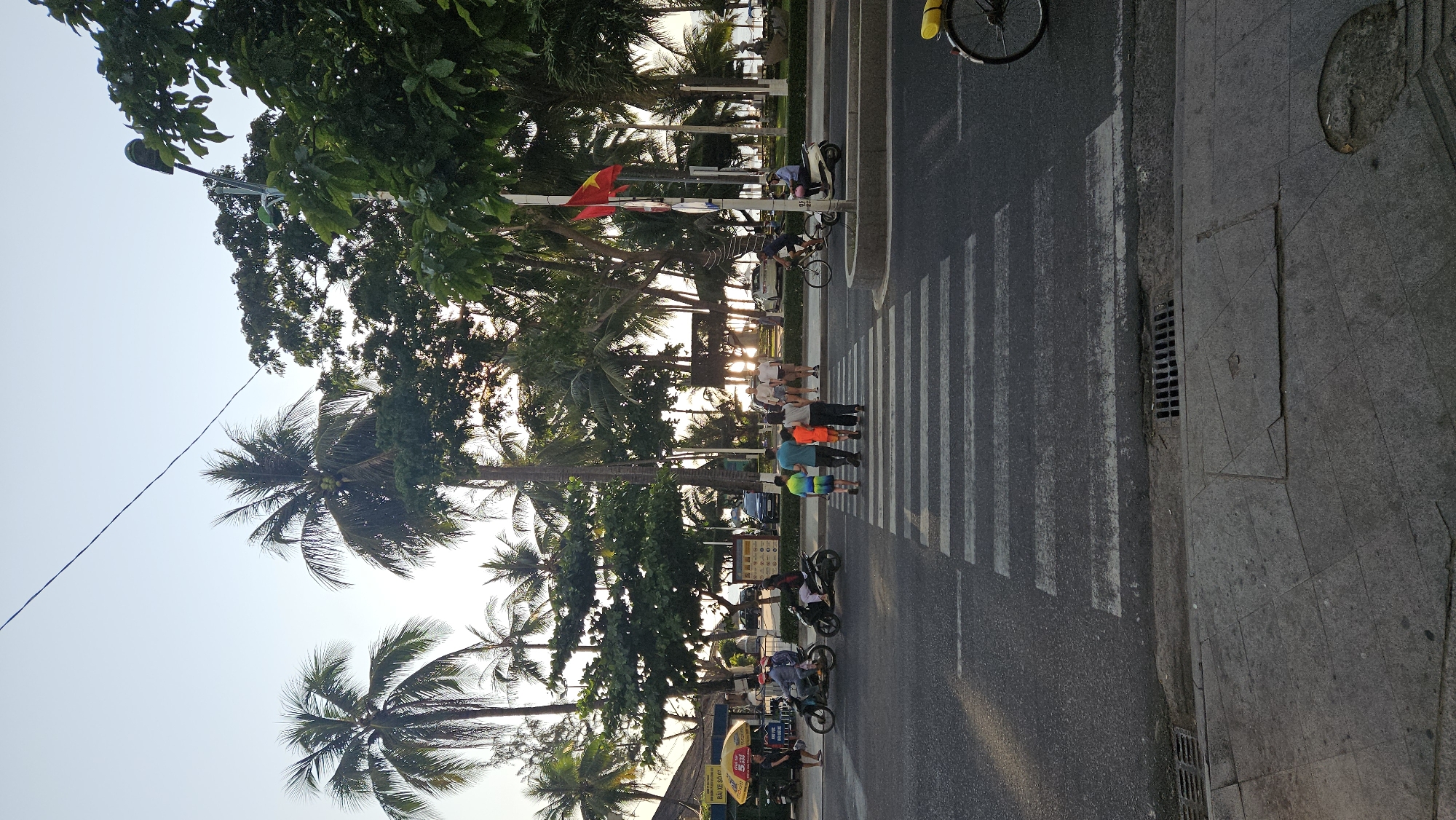 The image shows a street scene in Nha Trang, Vietnam. A group of people are crossing the street at a zebra crossing. Several motorbikes and bicycles are also present on the road. Palm trees line the street, and a Vietnamese flag is visible. The environment appears to be a coastal area with a clear sky. The image is taken during the day, with the sun possibly setting or rising, as there is a glow of light in the distance.