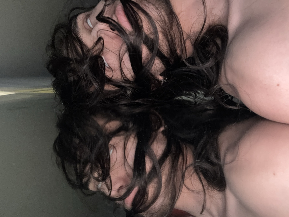 The image displays a horizontally mirrored depiction of a person, likely an adult, visible from the shoulders upward. The subject possesses long, dark, wavy hair that frames and partially obscures two mirrored partial faces. Both visible faces feature light-colored eyes rolled upwards, exposing a significant portion of the sclera, and slightly parted lips. Below the hair and faces, bare shoulders and upper chest are visible, indicating the individual is unclothed or partially clothed. The background is uniformly dark and indistinct, consistent with an indoor environment. No specific architectural details, landmarks, or environmental cues are present to determine a precise location within Sofia, Bulgaria.