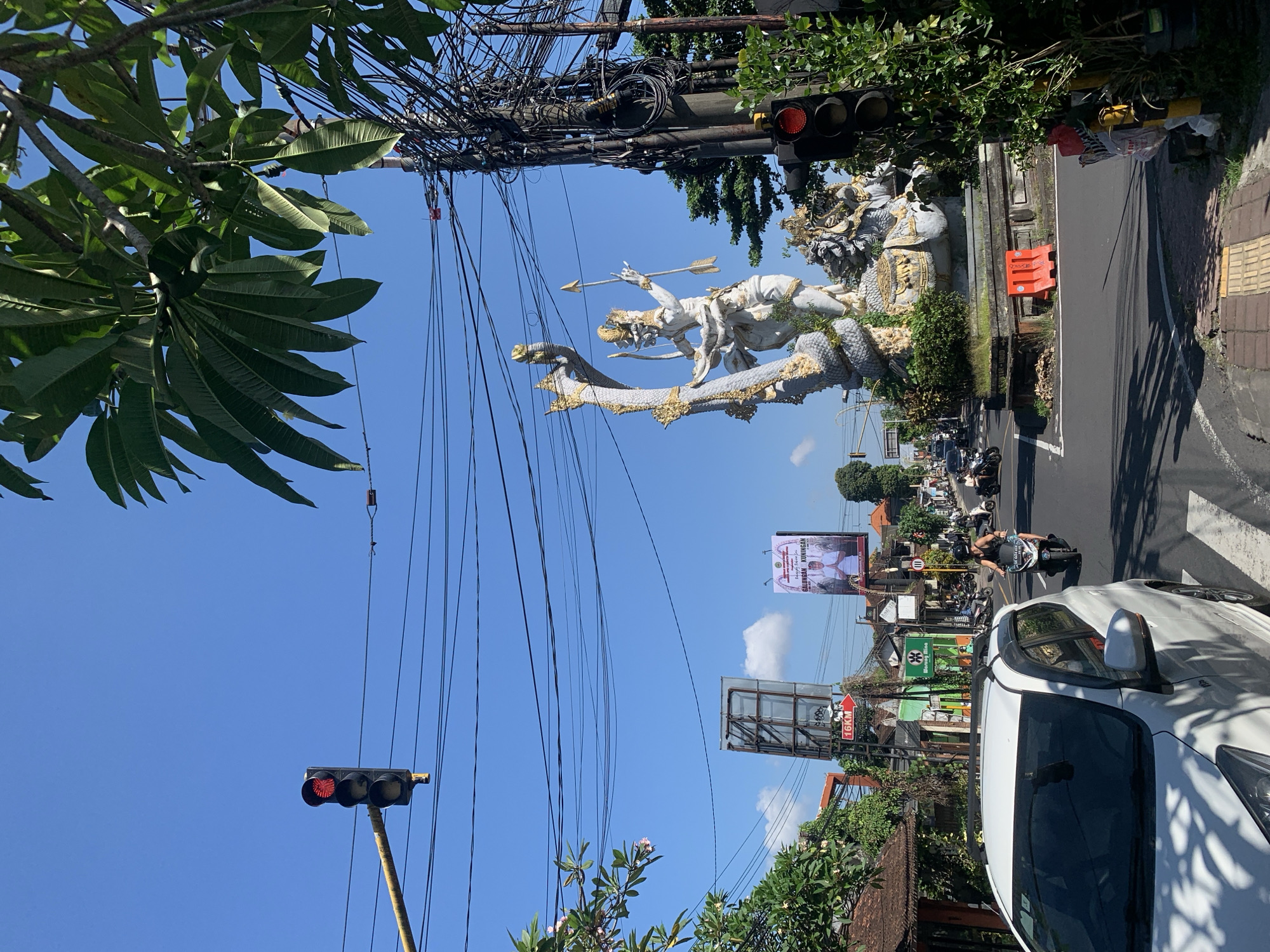 The image captures a street scene in Ubud District, Indonesia, characterized by a bright blue sky with scattered clouds. Prominently featured is a large, ornate statue depicting a mythical figure, possibly from Balinese mythology, intertwined with a serpent. The statue is white with gold accents and stands on a raised platform at a street intersection. To the left of the statue, a traffic light shows a red signal.
In the foreground, a white car is parked or driving on the left side of the road. The street is paved with asphalt and bordered by sidewalks with tactile paving. On the right side of the intersection, another traffic light is visible, also showing red. Overhead, a dense network of electrical wires crisscrosses the sky, supported by utility poles.
The surrounding environment includes lush green trees and vegetation, with some flowering plants. Buildings with traditional Balinese architectural elements are visible along the street, along with billboards. One billboard displays a man in formal attire with text in Indonesian. Traffic, including motorcycles and cars, moves through the intersection. A person is seen riding a motorcycle in the middle ground. The overall impression is a bustling urban environment with a strong cultural presence, typical of a tourist destination like Ubud.