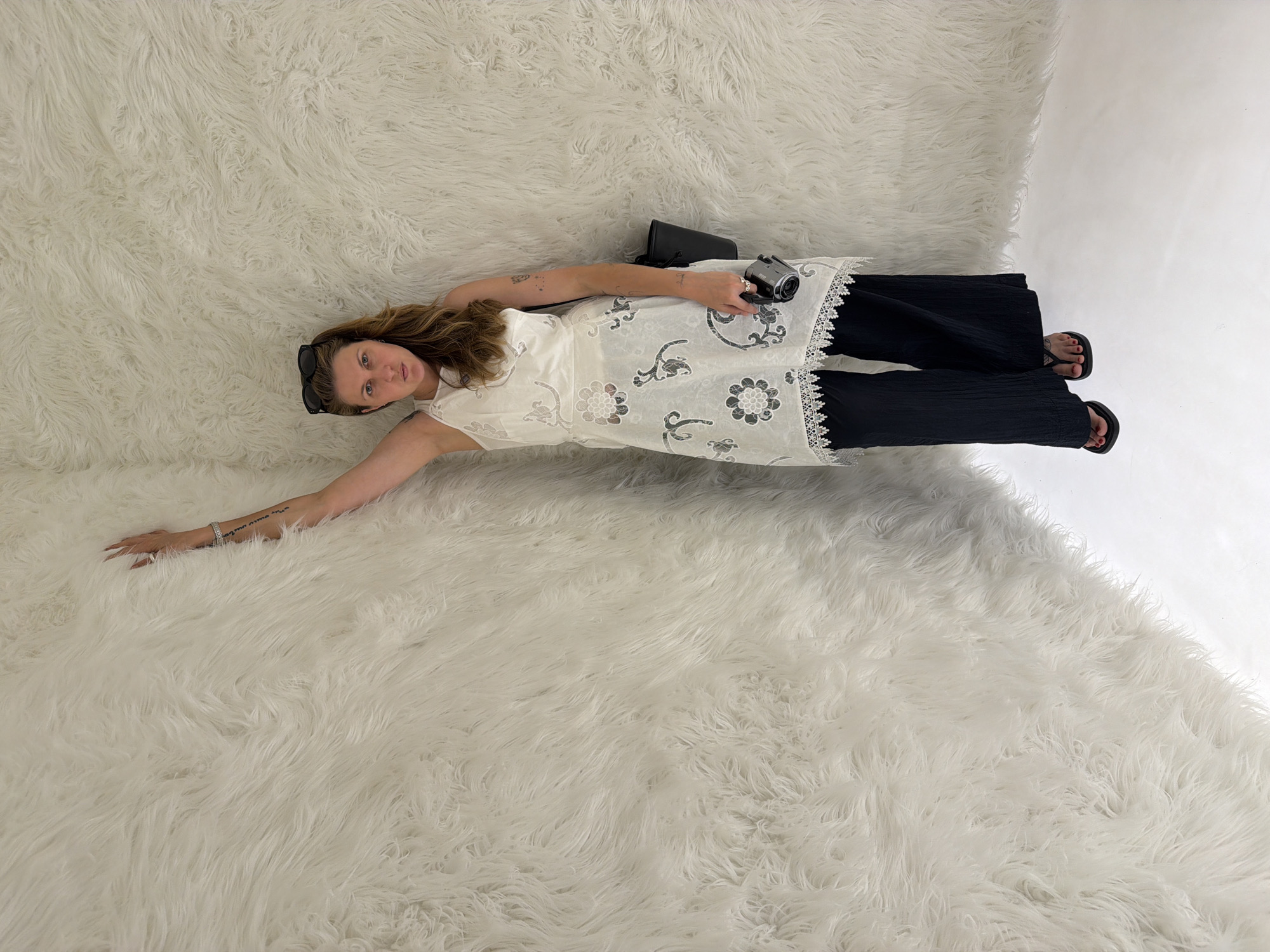 A woman with blonde hair stands in a corner with a white furry wall and white floor. She is wearing a white sleeveless dress with a floral pattern and black wide-leg pants. She has sunglasses on her head, a black bag over her shoulder, and is holding a camera in her right hand. Her left arm is extended upwards, with her hand resting on the furry wall. The scene is well-lit, and the woman is facing the camera. There is no indication of the location in Cold Spring, United States.