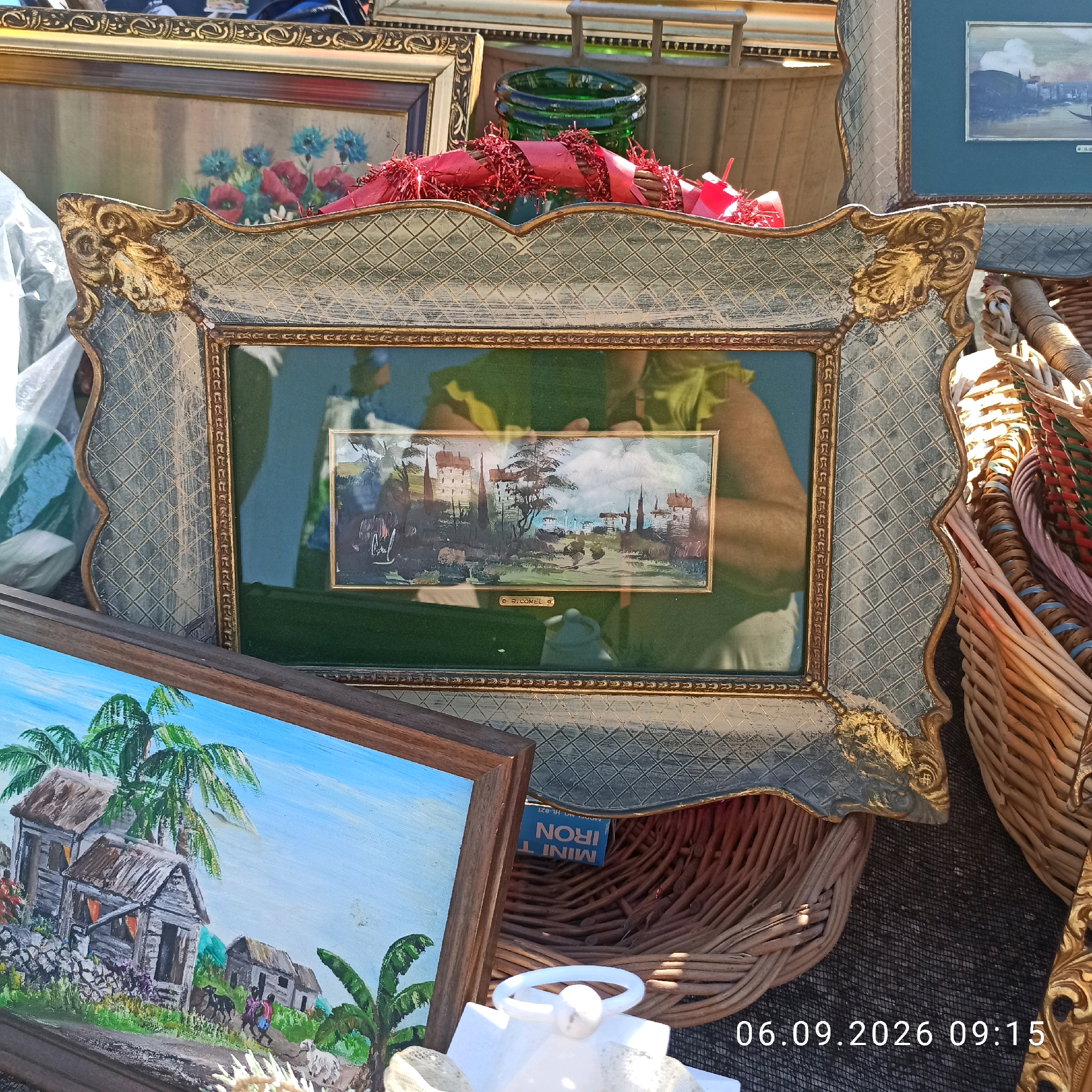 The image depicts a street market scene, possibly in Sannicandro di Bari, Italy. The foreground is dominated by several framed artworks and wicker baskets. A prominent, ornate, light-blue and gold-trimmed frame displays a small, horizontal landscape painting of a village. To the left, another framed painting shows a cluster of red and blue flowers. Below these, a rustic painting of tropical houses with palm trees is partially visible. Several wicker baskets, some filled with colorful yarn or fabric, are arranged around the art pieces. In the background, a green glass bottle and a tangle of red ribbon are visible, suggesting a festive or decorative element. A portion of a person's arm and shoulder, wearing a light-colored top, is reflected in the glass of the village painting. The ground appears to be covered in a dark mesh or netting. The date "06.09.2026 09:15" is superimposed on the lower right of the image.