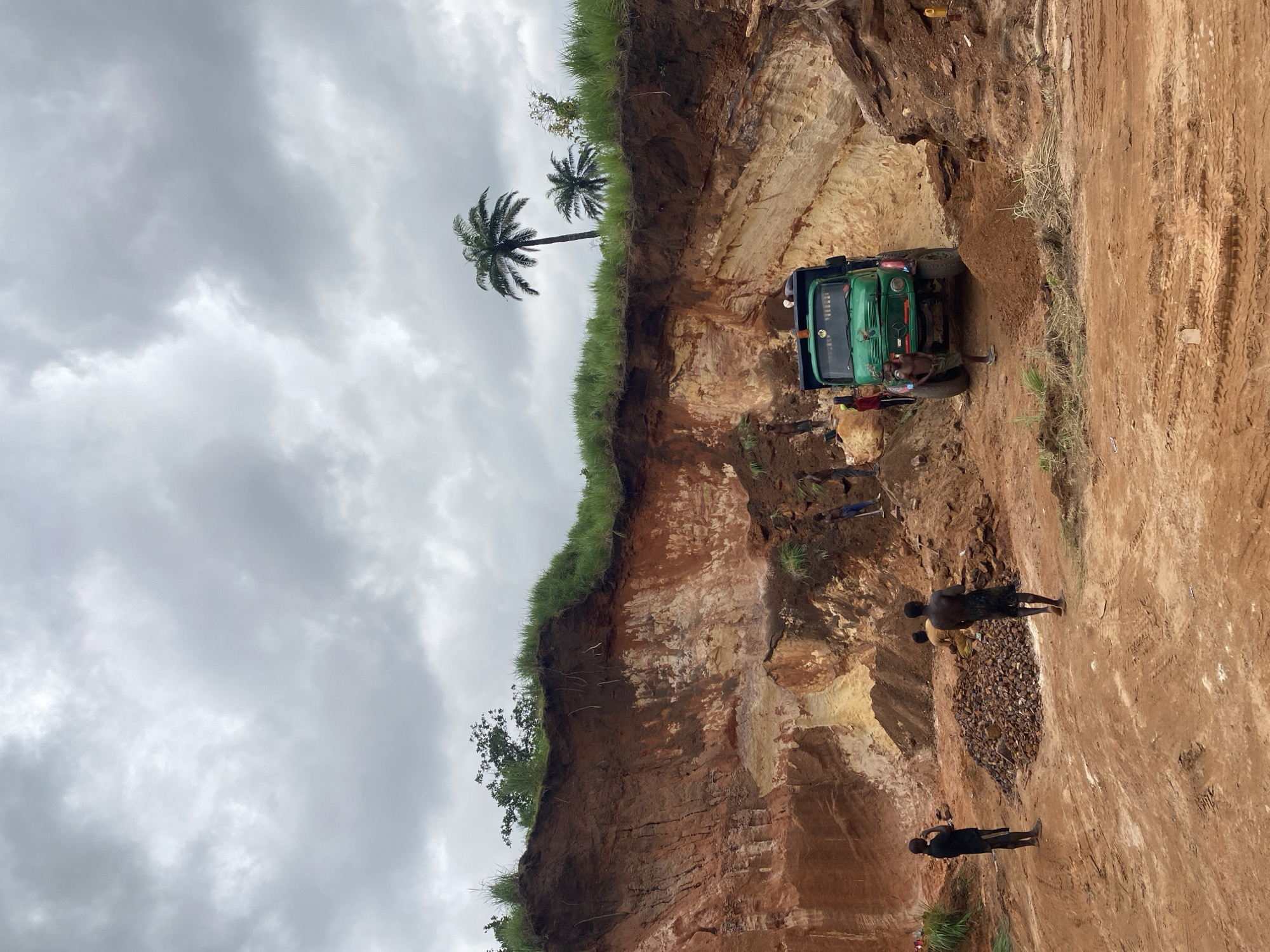 The image depicts an open-pit mine in Achara, Nigeria, under a cloudy sky. The mine face reveals layers of reddish-brown and yellowish earth, with a grassy slope on top. Several individuals are present, engaged in mining activities. A green dump truck is positioned at the base of the mine, appearing to be loaded or about to be loaded. Piles of excavated material are visible on the ground. In the foreground, a heap of rocks sits on the red earth. Several men, some appearing to be children, are scattered throughout the scene, with some standing and observing, while others are near the truck and the excavated material. Two palm trees are visible in the background, emerging from the top of the mine. The overall environment suggests an active, manual mining operation.