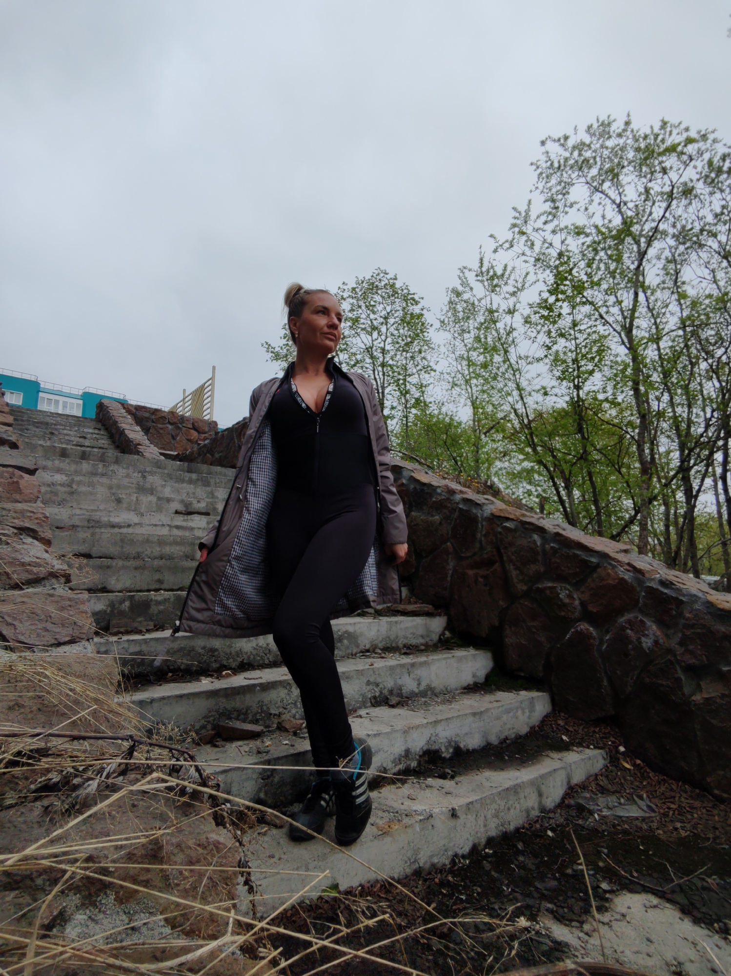 An adult Caucasian female stands on concrete stairs outdoors, looking to her right. She has blonde hair pulled up in a bun and is wearing a black jumpsuit and an unzipped grey jacket with a checkered lining. She has on athletic shoes. The stairs are bordered by a stone wall on her right and a building with blue siding and white windows in the background to her left. There are trees with green leaves visible behind the stone wall and in the distance. The sky is overcast and grey. The scene appears to be in the city of Petropavlovsk-Kamchatsky, Russia, given the context.
