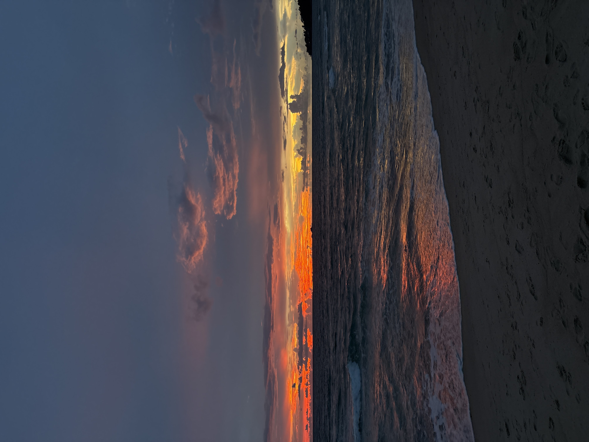 The image depicts a serene beach scene in Choeng Thale, Thailand, during twilight. The sky is a gradient of deep blue at the top, transitioning to vibrant oranges and reds near the horizon, indicating a sunset or sunrise. Wispy clouds are scattered across the sky, catching the colorful light.
The ocean is a dark, choppy expanse, reflecting the fiery hues of the sky. Gentle waves are breaking on the shore, creating a foamy white line. The beach in the foreground is composed of dark sand, with visible footprints scattered across its surface, suggesting recent human activity. To the far right, a dimly lit coastline with trees can be seen extending into the sea.
There are no people or specific objects clearly identifiable in the scene besides the natural elements of the beach, sea, and sky. The overall atmosphere is tranquil and natural, characteristic of a coastal landscape at dusk or dawn.
