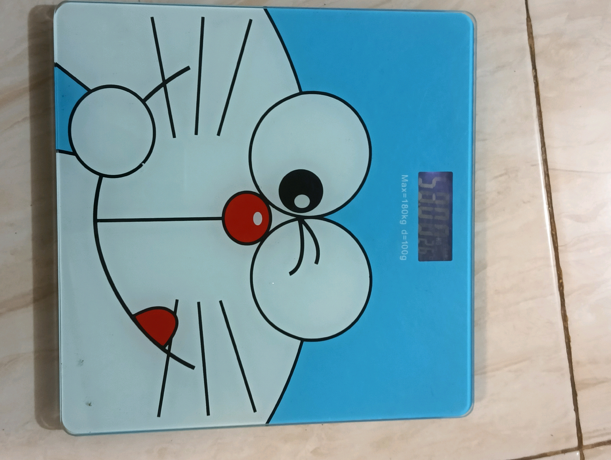 The image shows a blue digital bathroom scale with a cartoon character design, resembling Doraemon, on its surface. The scale displays a weight reading of "53.25". Text on the scale indicates "Max=180kg d=100g". The scale is placed on a tiled floor. The background suggests an indoor environment, likely a bathroom or bedroom, with light-colored tiles. There are no people or other significant objects in the immediate vicinity of the scale. The location is described as Banyuanyar Lor, Indonesia, but this information is not visually verifiable from the image itself.