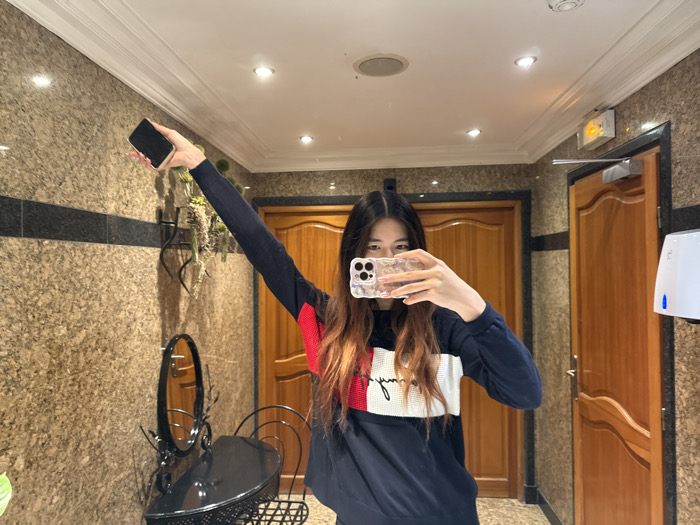 An image depicts a young Asian female taking a mirror selfie indoors within a facility located in Paris, France. She holds a smartphone with a clear, patterned case in her right hand, positioned to capture her reflection, obscuring her lower face. Her left arm is extended upwards, holding a second black smartphone. She is wearing a navy blue long-sleeved shirt with a prominent red, white, and navy horizontal stripe across the chest, featuring white text resembling a brand logo. The background consists of walls clad in speckled beige granite or marble with a horizontal black accent band. The white ceiling contains recessed lighting fixtures and a circular smoke detector. To the left, an ornate black metal vanity table with an oval mirror and a matching chair is positioned. A wall-mounted decorative planter with artificial greenery is visible. Two light brown wooden doors with molded panels are present in the background. Above the right door, an illuminated yellow "SORTIE DE SECOURS" (Emergency Exit) sign is mounted, alongside a white, wall-mounted hand dryer with blue indicators.