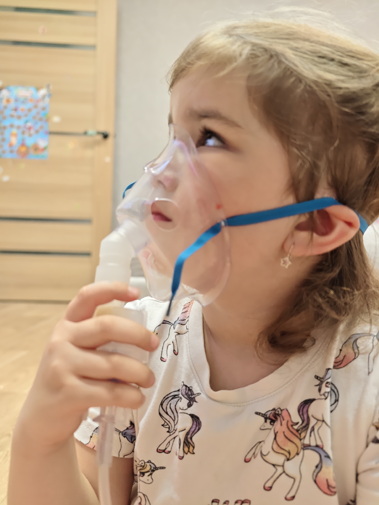 A young child is sitting indoors, using a nebulizer with a face mask attached. The child has light brown hair and is wearing a white t-shirt with a unicorn pattern. She is holding the nebulizer in her right hand and looking up and to the left. The mask is held in place with a blue strap. A door and a wall with a decorative picture or calendar are visible in the background. The location is indoors in the city of Chaikovsky, Russia. The flooring is light-colored wood laminate. The child is wearing a small, star-shaped earring in her left ear.