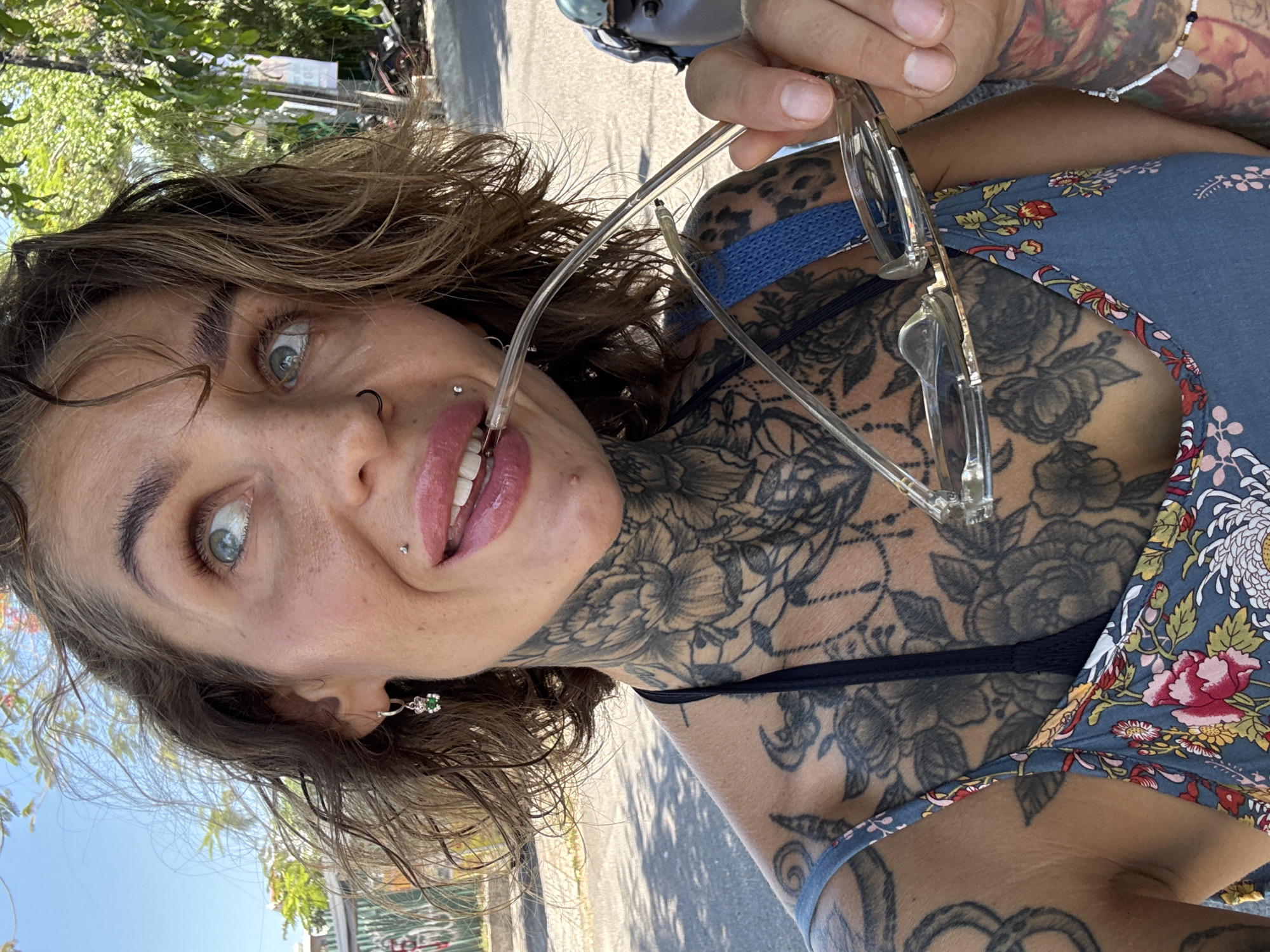 A woman with multiple tattoos on her neck, chest, and arm, and piercings on her nose and lip, is holding a pair of eyeglasses up to her mouth. She has blue eyes, brown hair, and is wearing a floral dress. She is outdoors, with a street and trees visible in the background. The location is Bac Nha Trang Ward, Vietnam.