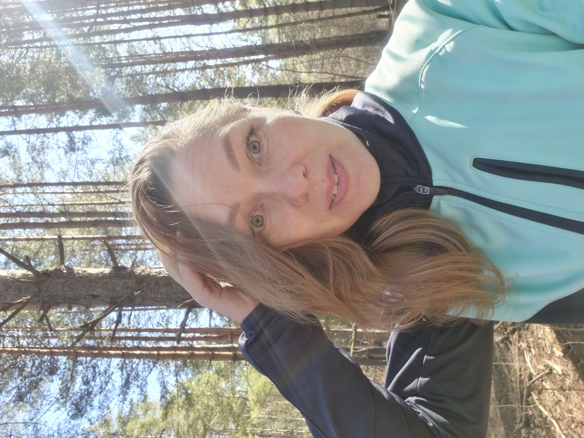 A light-skinned woman with light brown hair and green eyes is the central subject. She is wearing a light blue jacket with dark navy blue trim. She is holding her hand up near her hair, with a possible forest area in the background. The location cannot be determined from the image. There is no evidence to indicate this image was taken in Chaikovsky, Russia.