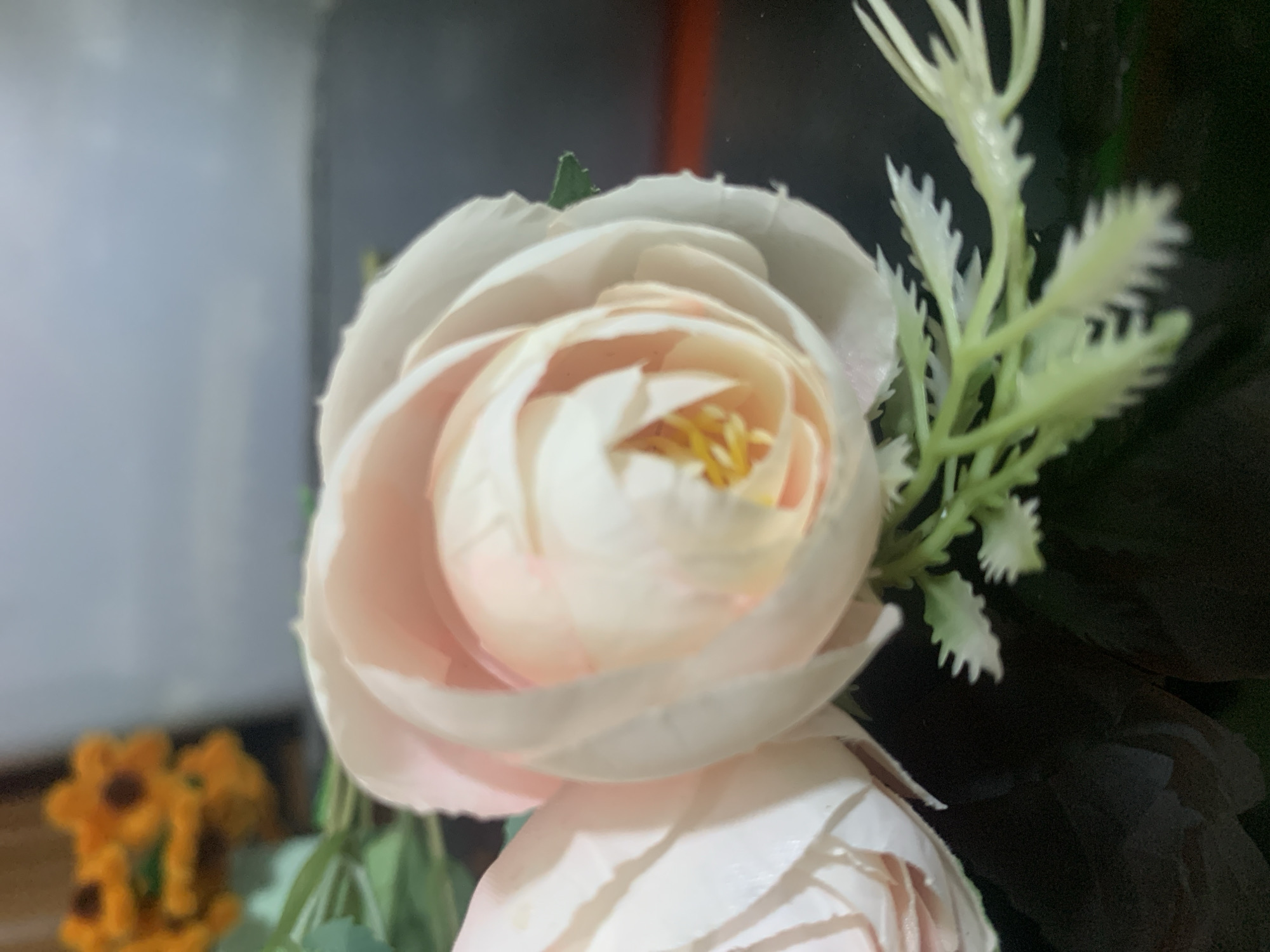 The image shows a close-up of two artificial light pink roses with yellow centers. The roses are on a dark, reflective surface. In the background, out of focus, are some artificial yellow flowers that resemble sunflowers. The scene is likely an indoor setting, possibly a decorative arrangement. There are no people or animals present. The image does not provide any specific information about the city of Kuala Pembuang, Indonesia.