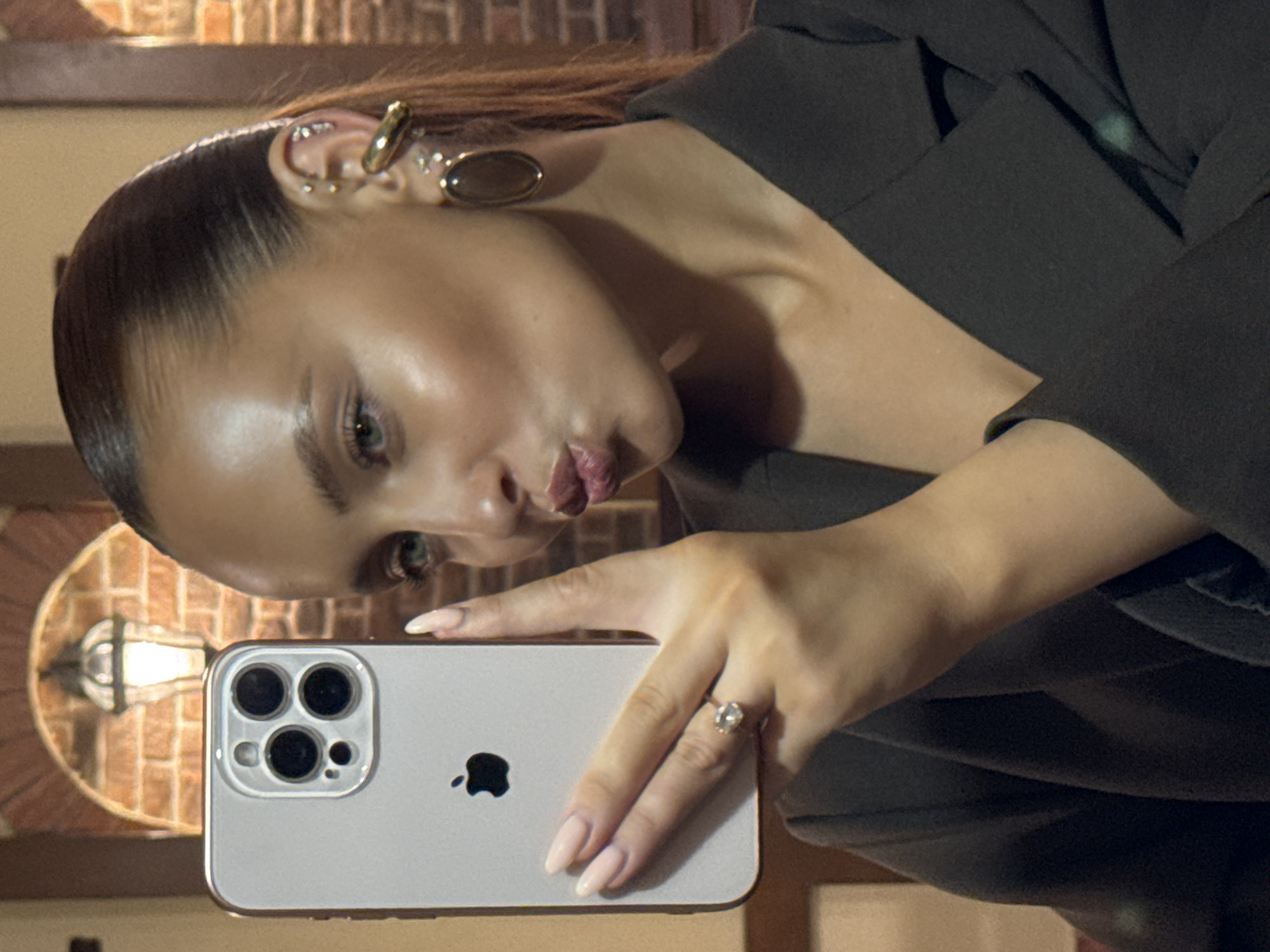 A woman with her hair pulled back is taking a selfie with a silver iPhone 13 Pro. She is wearing a black blazer and large earrings. The background appears to be a dimly lit restaurant with exposed brick and a lantern. The woman is pouting her lips towards the camera.