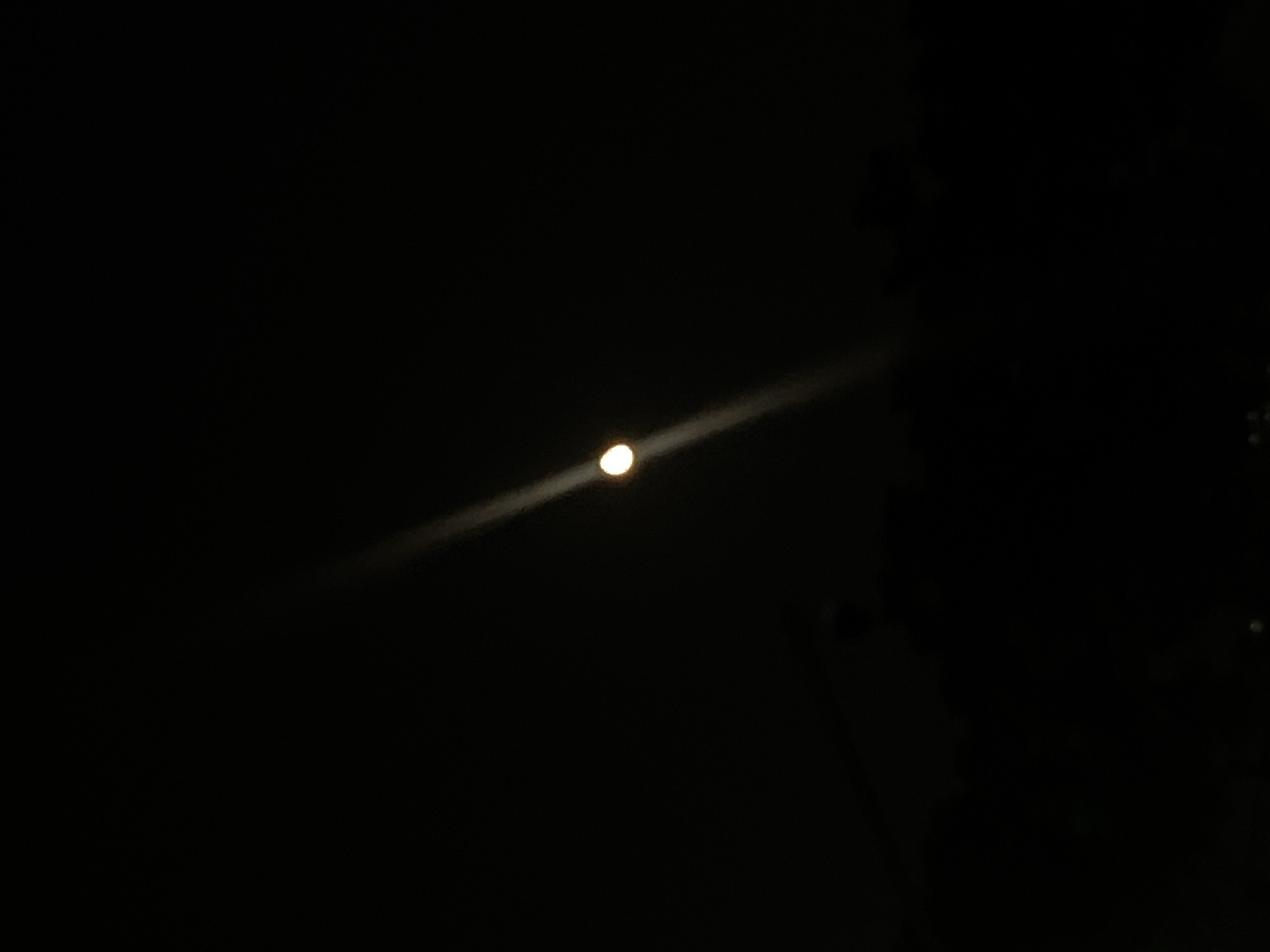 This forensic description details an image taken at night in Jiulongpo District, China. The scene is dominated by a profound darkness, characteristic of an unlit nighttime environment. A singular, intensely bright, circular light source, consistent with a celestial body such as the moon, is centrally positioned in the upper half of the frame. Originating from and extending diagonally across the frame, particularly towards the lower right, is a prominent, linear light streak. This streak, likely an optical aberration or lens flare, emanates directly from the primary light source. The lower quarter of the image presents a horizontal, amorphous dark mass, signifying a silhouetted landscape or distant urban elements against the night sky, with no discernible details. No individuals, animals, specific structures, or discernible actions are present within the frame.