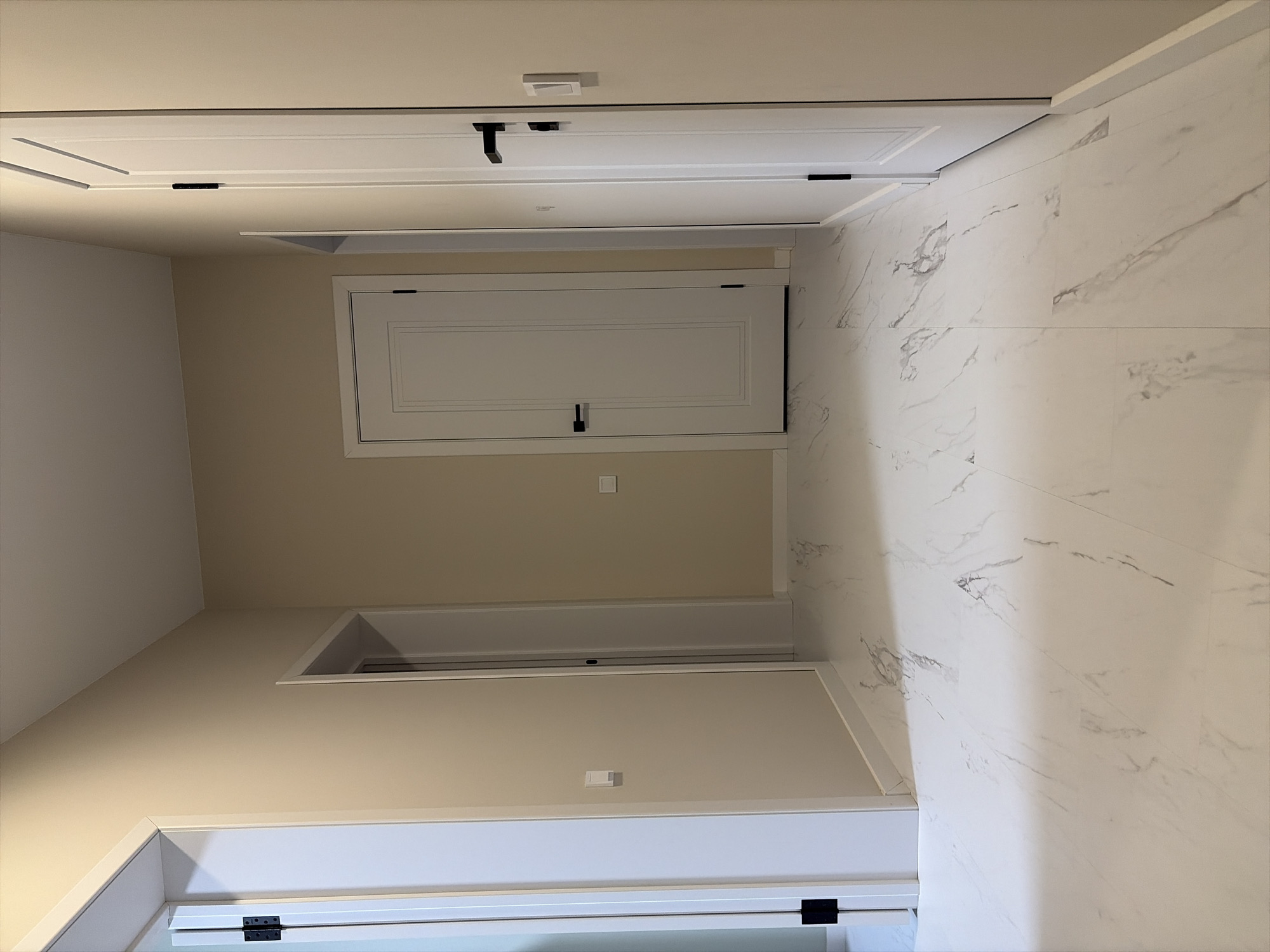 The image captures an empty interior hallway or corridor, viewed from a slightly elevated perspective. The space is well-lit, highlighting its modern and clean architectural design.
On the left side of the frame, a wall painted in a uniform light beige or tan color features multiple white doors with contrasting black hardware. At the top, a closed standard-sized white door is equipped with a contemporary black handle/latch mechanism. A small, square white object, possibly a sensor or light switch, is mounted on the wall directly above this door. Below it, on the beige wall, a smaller, horizontal white panel door, likely an access panel or compact storage, is also fitted with black hardware. A discreet white square switch or sensor is visible on the beige wall beneath this panel. Further down, an open doorway leads into another area, which appears to be painted in the same light beige hue. A partial view of another white door with black hinges is visible at the very bottom.
The right side of the hallway, including both the wall and the floor, is adorned with large, rectangular white tiles featuring prominent gray marbling patterns, creating a continuous and elegant surface. The flooring seamlessly matches the wall tiling. The overall aesthetic emphasizes a minimalist and contemporary style with a consistent palette of white, beige, and black accents. There are no people, visible activities, or discernible text in the image. The time of day and external conditions cannot be determined from this interior shot.