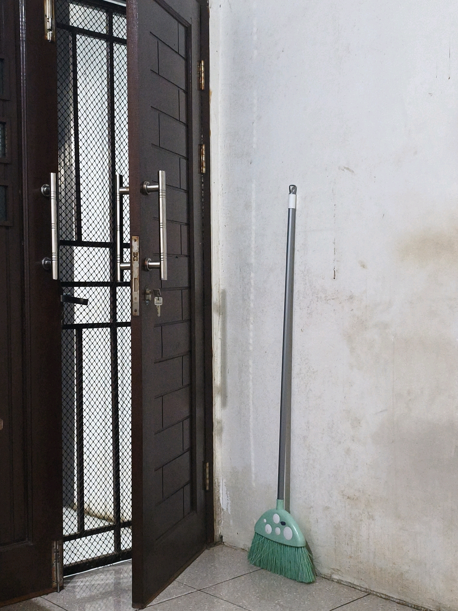 The image depicts an indoor scene, likely a hallway or entryway, with a focus on a dark brown wooden double door, one leaf of which is ajar. The doors feature metallic handles and a decorative patterned inlay. To the left of the doors, a metal security screen is visible. A broom with a green plastic head and white polka dot accents, and a gray handle, leans against a light-colored, somewhat grimy wall on the right side of the frame. The floor is tiled. The scene is devoid of people. The presence of the broom suggests an area that is or has been undergoing cleaning. The overall impression is that of a residential or functional space within Samarinda, Indonesia.