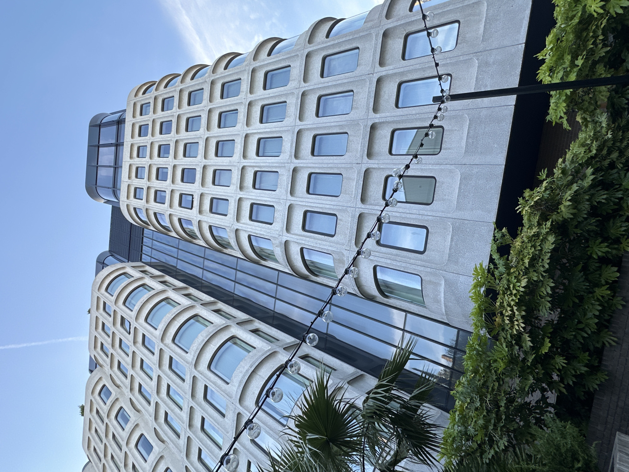 The image captures a low-angle view of a distinctive architectural building in London, United Kingdom. The structure features rounded, tiered sections with a grid-like facade of recessed windows. A contrasting dark glass section connects the two rounded tiers. A string of decorative lights hangs in the foreground, partially obscured by palm and other green vegetation. The sky is a clear, pale blue with a faint contrail visible.