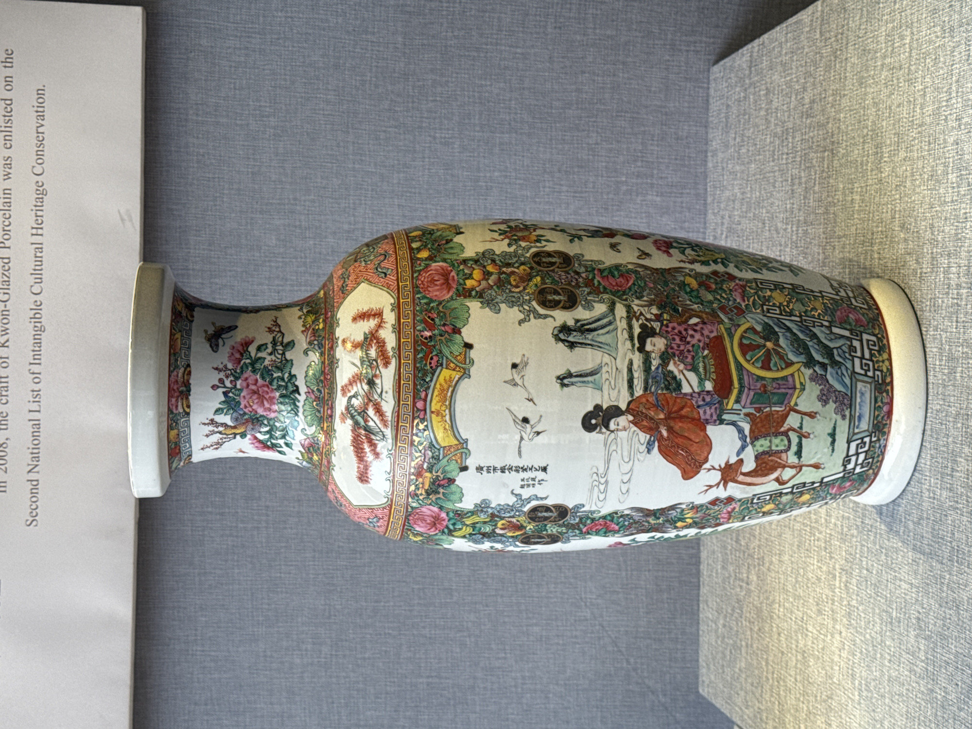 The image shows a Chinese porcelain vase with intricate floral and figural designs. The vase features a flared neck and a rounded body, decorated with colorful scenes of women in traditional robes riding in a cart pulled by deer, set against a backdrop of stylized clouds, cranes, and ornamental foliage. The vase also has a rectangular panel inscribed with Chinese characters, identifying it as “Canton Famille Rose Porcelain Craft Version, Liwan District, Guangzhou City”. The artifact appears to be displayed in a museum or gallery setting, with a textual description of the vase’s historical significance partially visible on the left side of the image.