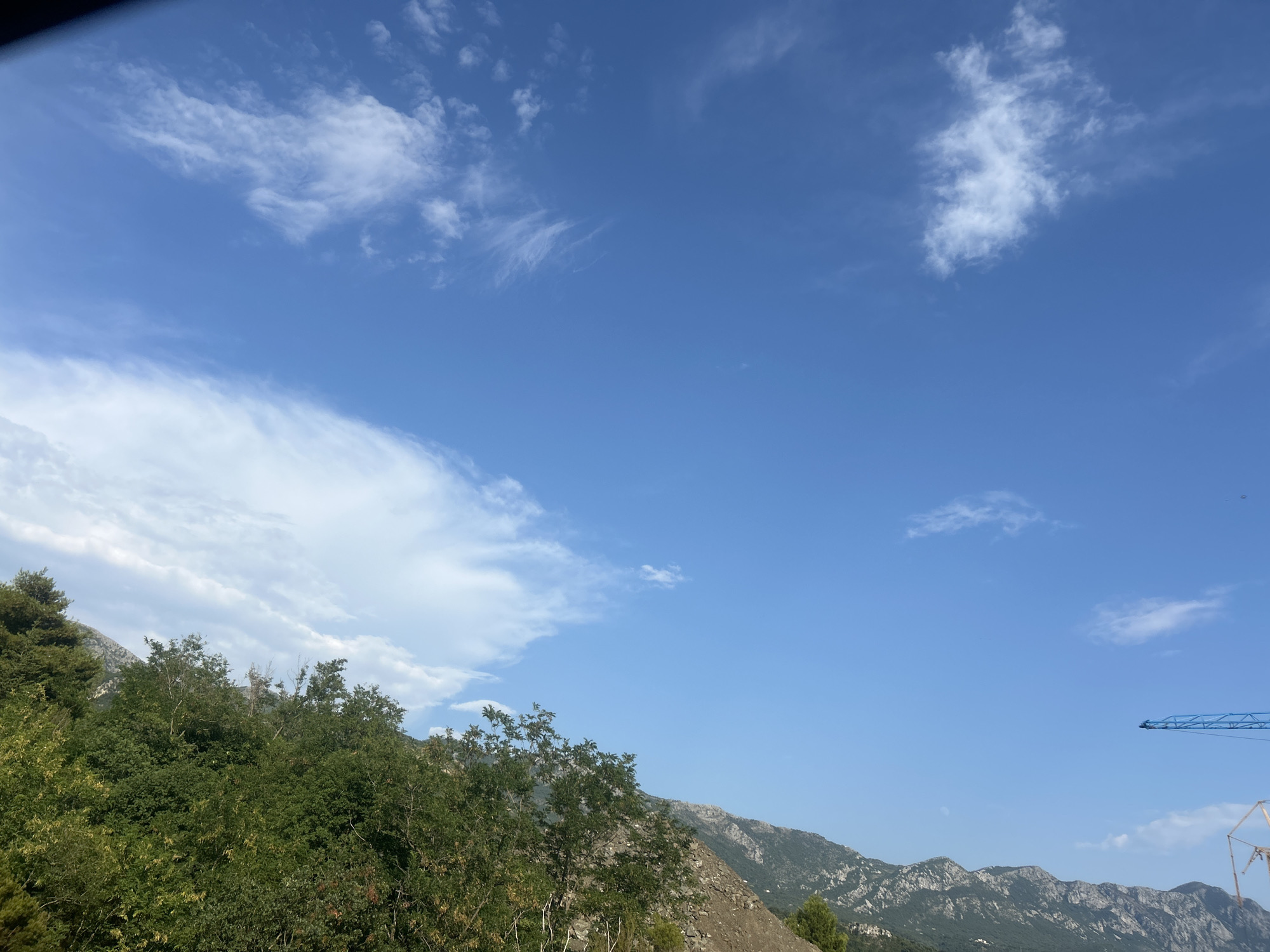 The image depicts a daytime outdoor scene, likely in Boreti, Montenegro, characterized by a vast blue sky populated with scattered white clouds of varying densities. The lower portion of the frame is dominated by lush green foliage, including trees and shrubs, that forms a dense canopy. Beyond the foliage, a rugged mountainous terrain stretches across the horizon, with visible rocky slopes and vegetation. In the far right of the image, a portion of a construction crane, colored blue, is visible against the sky, suggesting ongoing development or infrastructure work in the vicinity. The overall impression is one of a natural landscape juxtaposed with human construction activity.