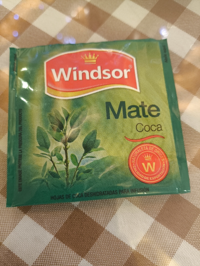 A sealed green packet of "Windsor Mate Coca" tea lies on a checkered surface. The packaging features a red logo with a golden crown atop the word "Windsor." Below, "Mate Coca" is printed in bold. A stylized coca plant image is on the left. A red circular stamp with "NATURALEZA DE ORIGEN" and "CALIDAD DE EXPORTACION" surrounds a letter "W" with a crown. Text at the bottom reads "HOJAS DE COCA DESHIDRATADAS PARA INFUSION," which translates to "DEHYDRATED COCA LEAVES FOR INFUSION". "ESTE ENVASE PROTEGE LA FRESCURA DEL PRODUCTO" is partially visible and means "THIS PACKAGE PROTECTS THE FRESHNESS OF THE PRODUCT."