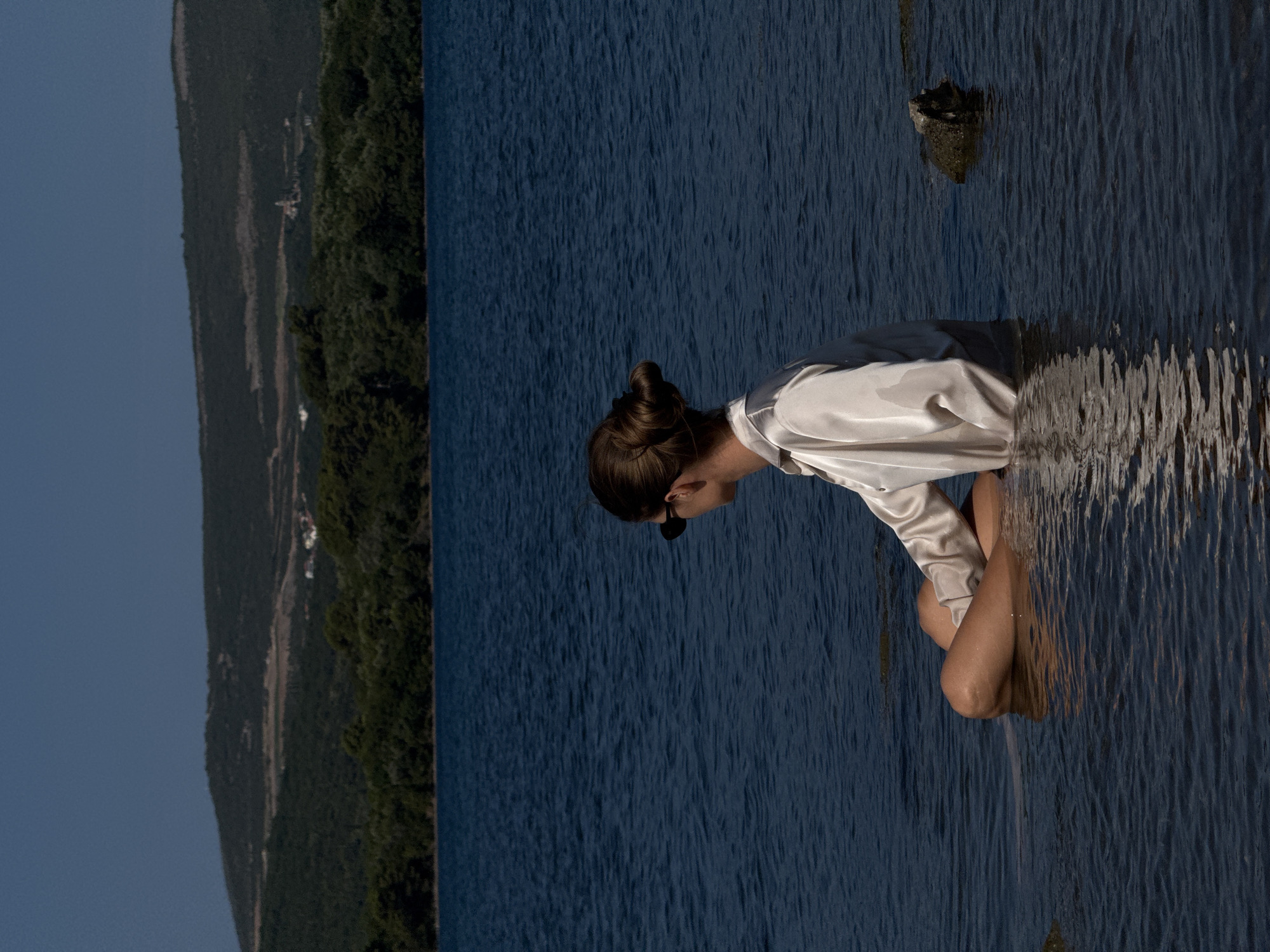 A woman sits cross-legged in shallow water, facing away from the camera. She has her hair styled in a bun and wears sunglasses. She is dressed in a light-colored, possibly silk, button-up shirt. The water is a deep blue, with ripples reflecting the sky and the woman. In the background, a lush green treeline separates the water from a hilly landscape under a clear blue sky. The location is identified as Mrčevac, Montenegro. A large rock is visible to the right of the woman in the water.