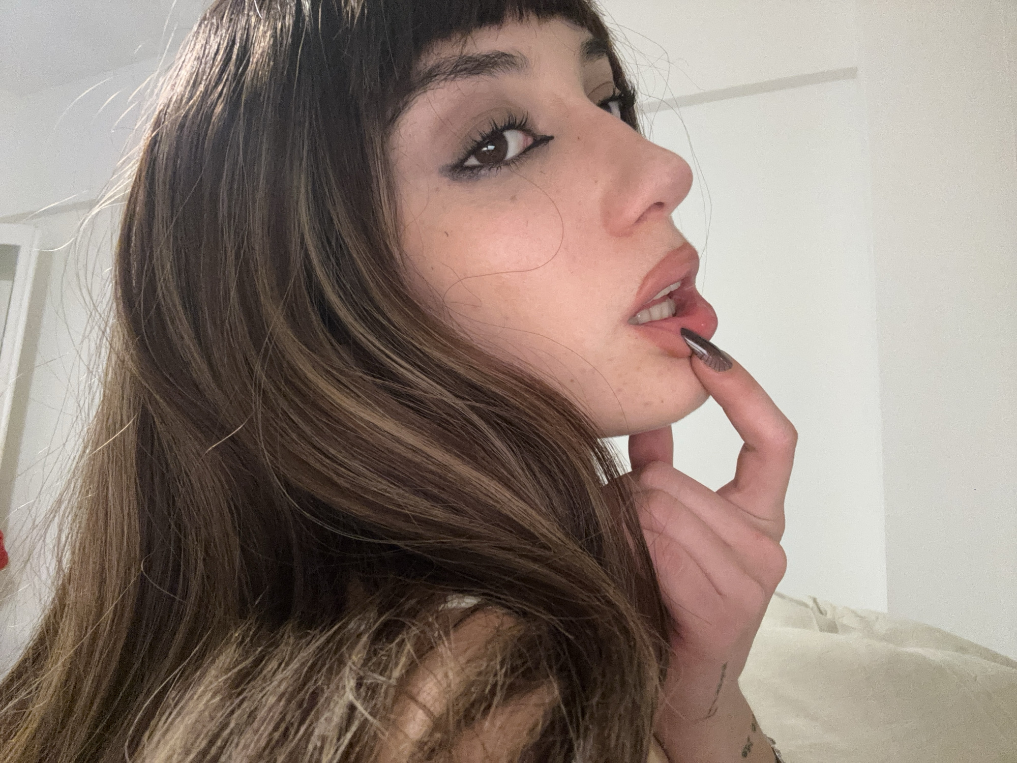 A young woman with brown hair, styled with bangs, is positioned indoors. She has fair skin and is wearing makeup, including eyeliner. Her finger is touching her lips. The background shows a white wall and a glimpse of a doorway. The location appears to be an interior space, but there is no indication to confirm if the location is within Buenos Aires, Argentina.