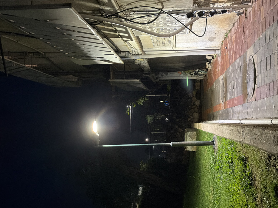This image depicts an outdoor urban pathway at night in Migdal HaEmeq, Israel. A tall, greenish-blue metallic lamppost with a single luminaire stands prominently on the left, emitting bright light that illuminates the immediate foreground. To the left of the lamppost is a manicured grassy area. A concrete curb separates this grass from a brick-paved pathway on the right. The pathway is constructed from interlocking rectangular bricks, primarily red and gray, and appears damp. Two circular metal manhole covers are embedded in the pathway.
To the right of the pathway, a light-colored building wall rises vertically, adorned with numerous dark cables, white conduits, and electrical boxes. A window with a white metal security grate is visible on this wall. Above the window, a large, slatted or louvred architectural element extends horizontally.
In the mid-ground, beyond the pathway and behind the lamppost, is a low stone or rock retaining wall topped with dense dark vegetation. Further in the distance, approximately two blurry figures are discernible on an unpaved area or path, and another distant light source is visible. The sky overhead is dark. No specific actions or interactions are clearly discernible among the distant figures.