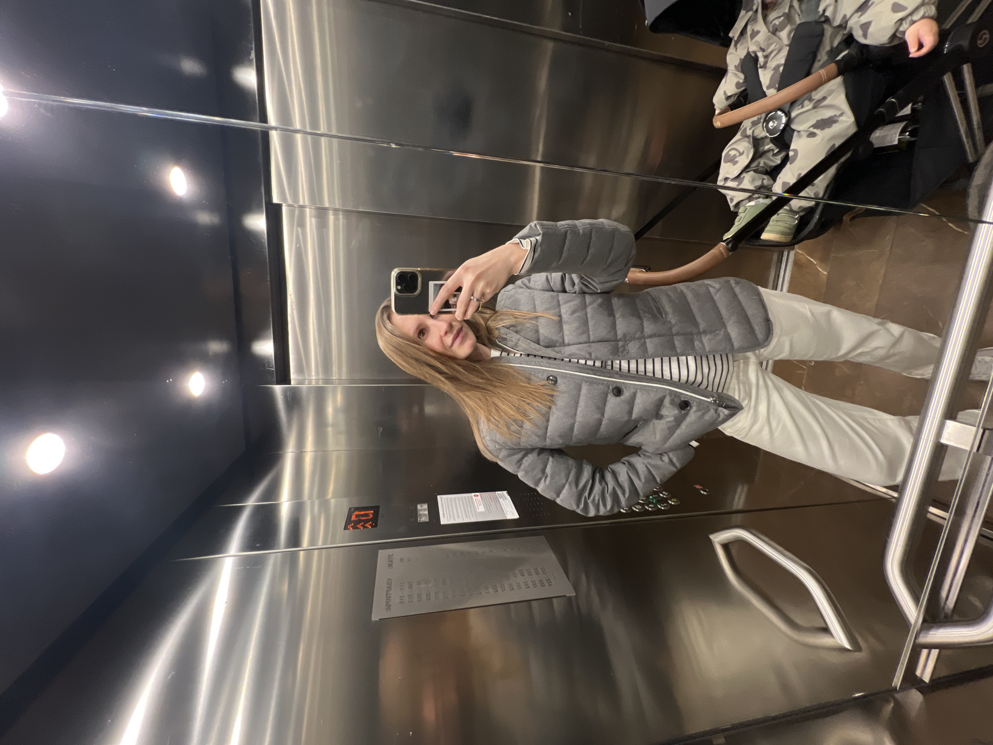 A full-length mirror selfie captures a young woman and a child in an elevator with brushed stainless steel walls and a dark floor. The woman, with long blonde hair, stands on the left side of the frame, holding a smartphone with a dark case in her right hand to take the photo. She is wearing a grey quilted jacket over a black and white striped top and white pants. A silver ring is visible on her left ring finger. She has a slight, pleasant smile.
Reflected on the right side of the frame, behind the woman, is a child seated in a dark stroller or pushchair. The child is dressed in a grey and white camouflage-patterned jumpsuit and green sneakers, secured by a dark harness and a brown safety bar.
The elevator interior features bright overhead lights, which are reflected in the shiny metallic surfaces. On the elevator control panel, a digital display shows "4U" (possibly floor 4) in orange. Below it, a directory panel provides various numbered sequences, likely room or floor indicators, and text in Russian, including "КЕМПИНГ" (KEMPING), meaning "camping." A smaller white sticker with illegible text is also present. The scene is indoors, well-lit by artificial lights, and located in Moscow, Russia.