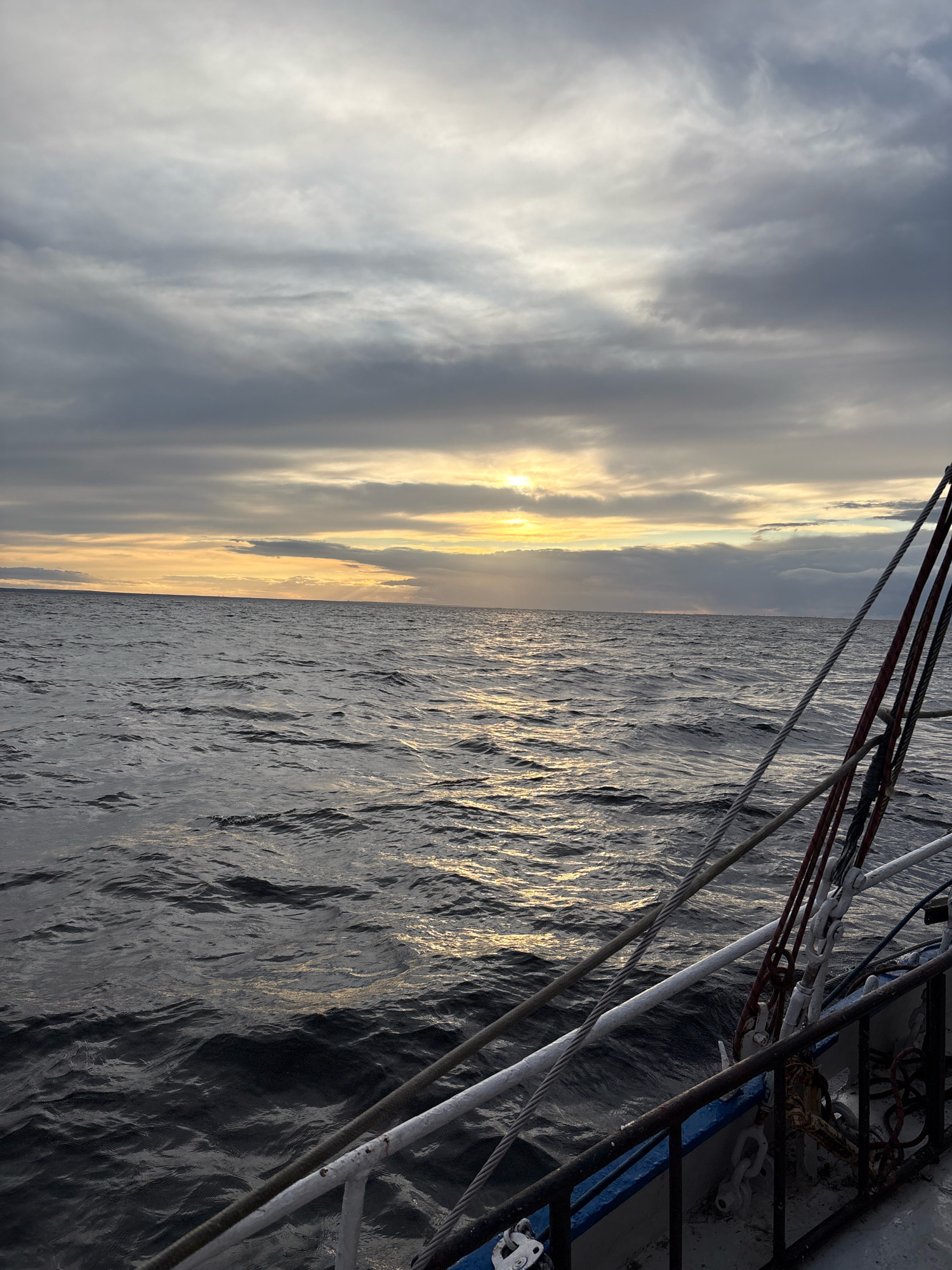 The image depicts a view from a boat sailing on a body of water. The sky is overcast with clouds, but a sliver of sunlight is visible breaking through near the horizon, casting a warm glow on the water's surface. The sea appears choppy with visible waves. In the foreground, the edge of the boat is visible, with rigging and cables present. The boat has a railing and appears to be a vessel designed for sailing. There are no people visible in the image. The scene is likely taking place at sea, possibly in the Gulf of Finland, given the mention of Saint Petersburg, Russia, as the location.