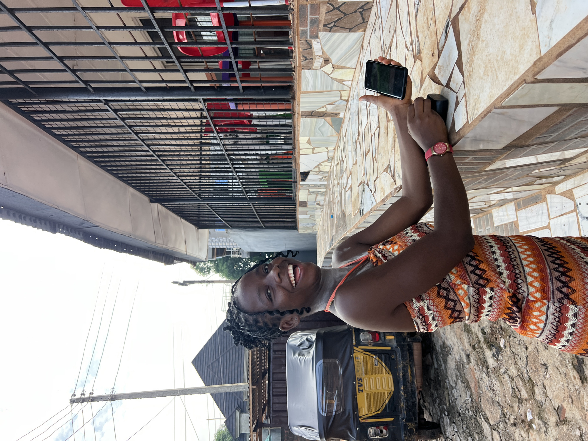 A young Black woman is taking a selfie with her smartphone. She is smiling broadly and her head is tilted towards the camera. She is wearing a colorful patterned dress and a red watch. The background shows a street scene in Nkpor, Nigeria, with a tiled wall, a metal grate, and a yellow auto-rickshaw. The sky is bright, suggesting it is daytime, and the weather appears to be clear. There is a sign on the auto-rickshaw that says "TVS".