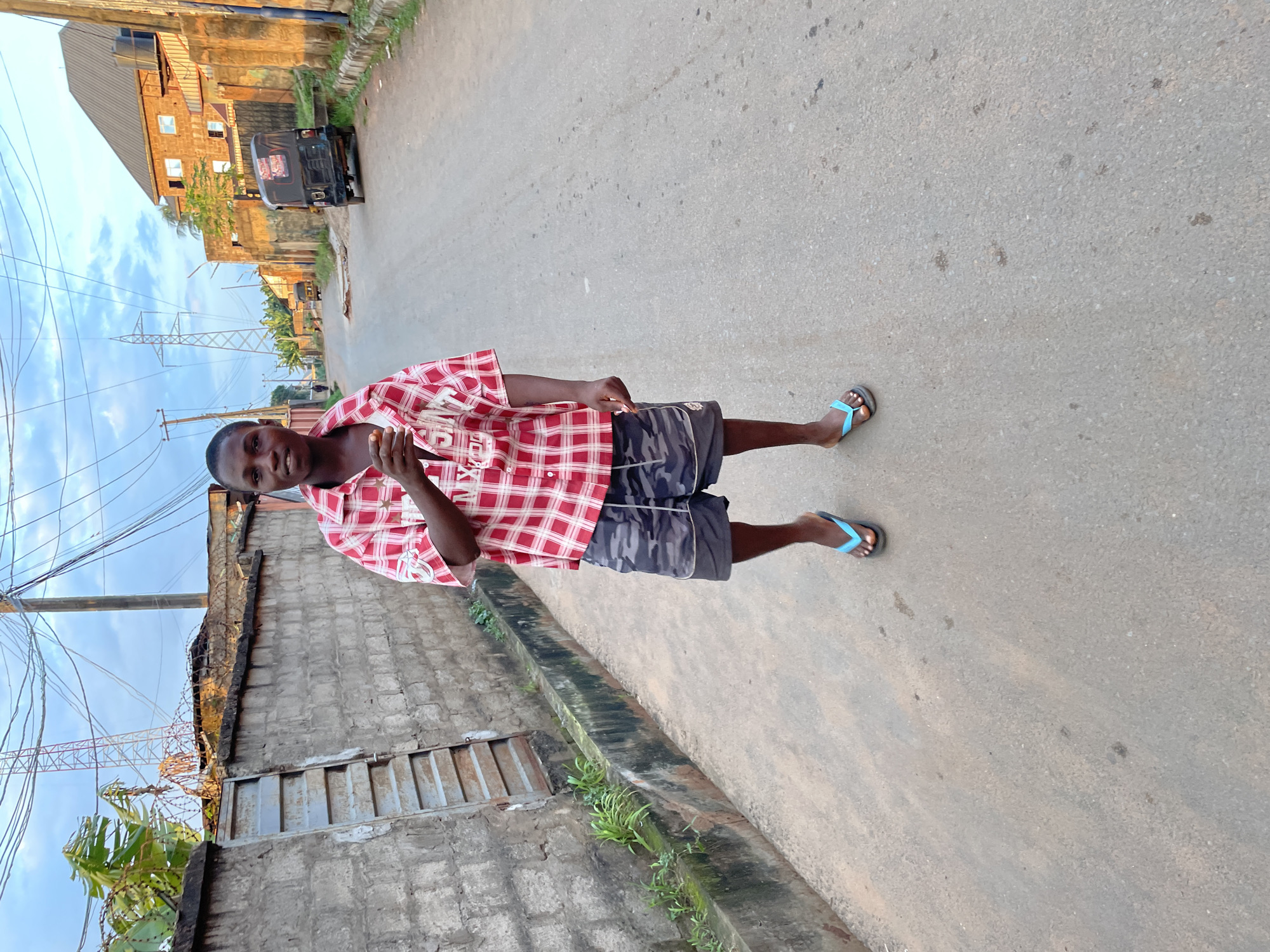 In Nkpor, Nigeria, a young male stands on a paved road. He is wearing a red and white checkered short-sleeved shirt, camouflage shorts, and blue flip-flops. The road is lined with a concrete wall on the left, which has some greenery growing along its base. On the right side of the road, there are buildings and a three-wheeled vehicle. The sky is visible in the background, with some clouds and power lines stretching across it. A tall communication tower is also present in the distance. The lighting suggests it is late afternoon or early evening.