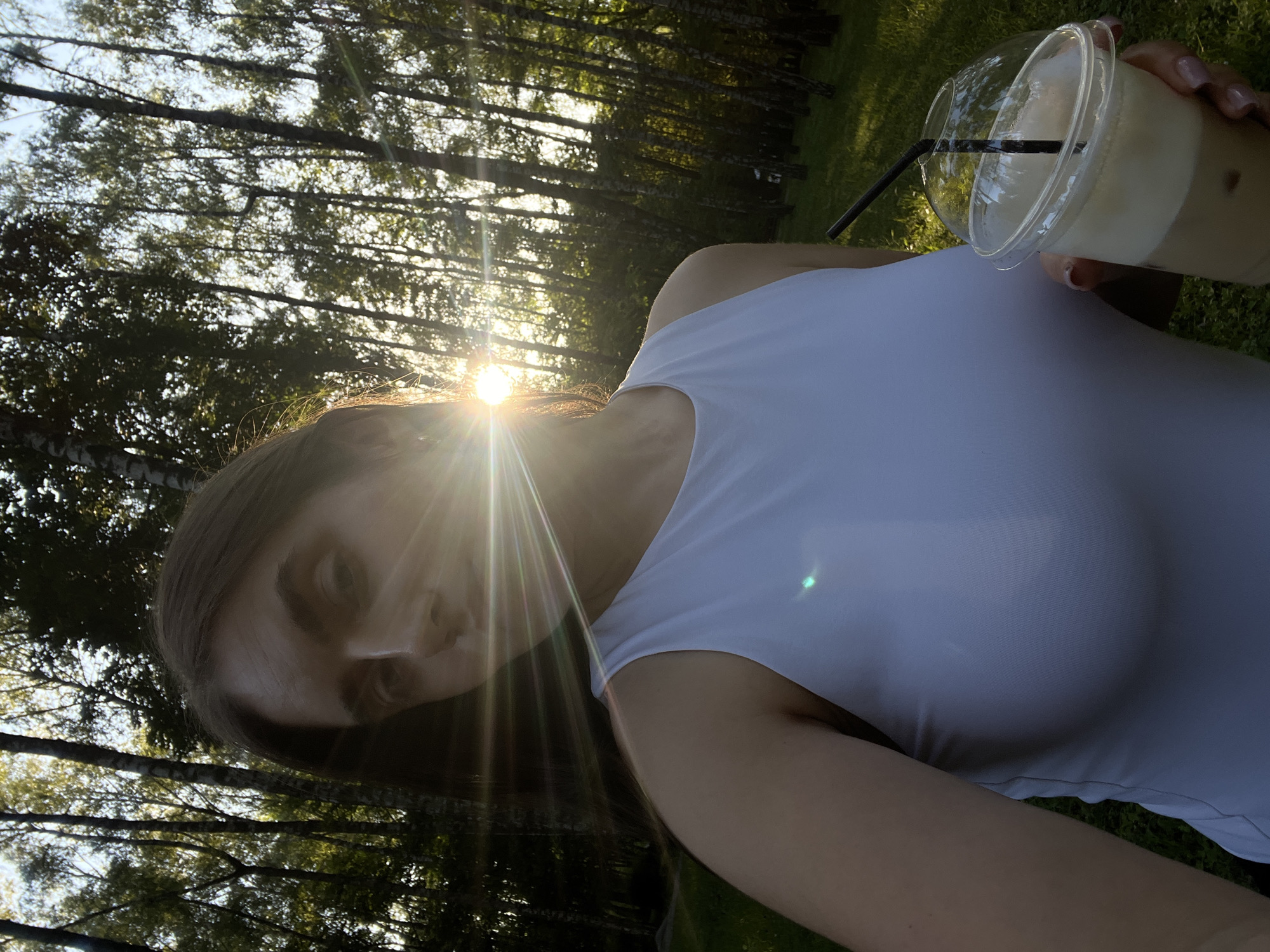 The image captures a close-up selfie taken outdoors, with a person holding a clear plastic cup filled with a light-colored beverage and a black straw. The person is wearing a white tank top. The background consists of a dense forest with tall trees, and the sun is setting, casting a warm glow and lens flares through the branches. The light suggests it is late afternoon or early evening. The setting appears to be a grassy area at the edge of the woods. The overall mood is relaxed and natural, possibly indicating leisure time. The image is oriented vertically. There is no visible text in the image. The location is identified as Ternopil, Ukraine.
