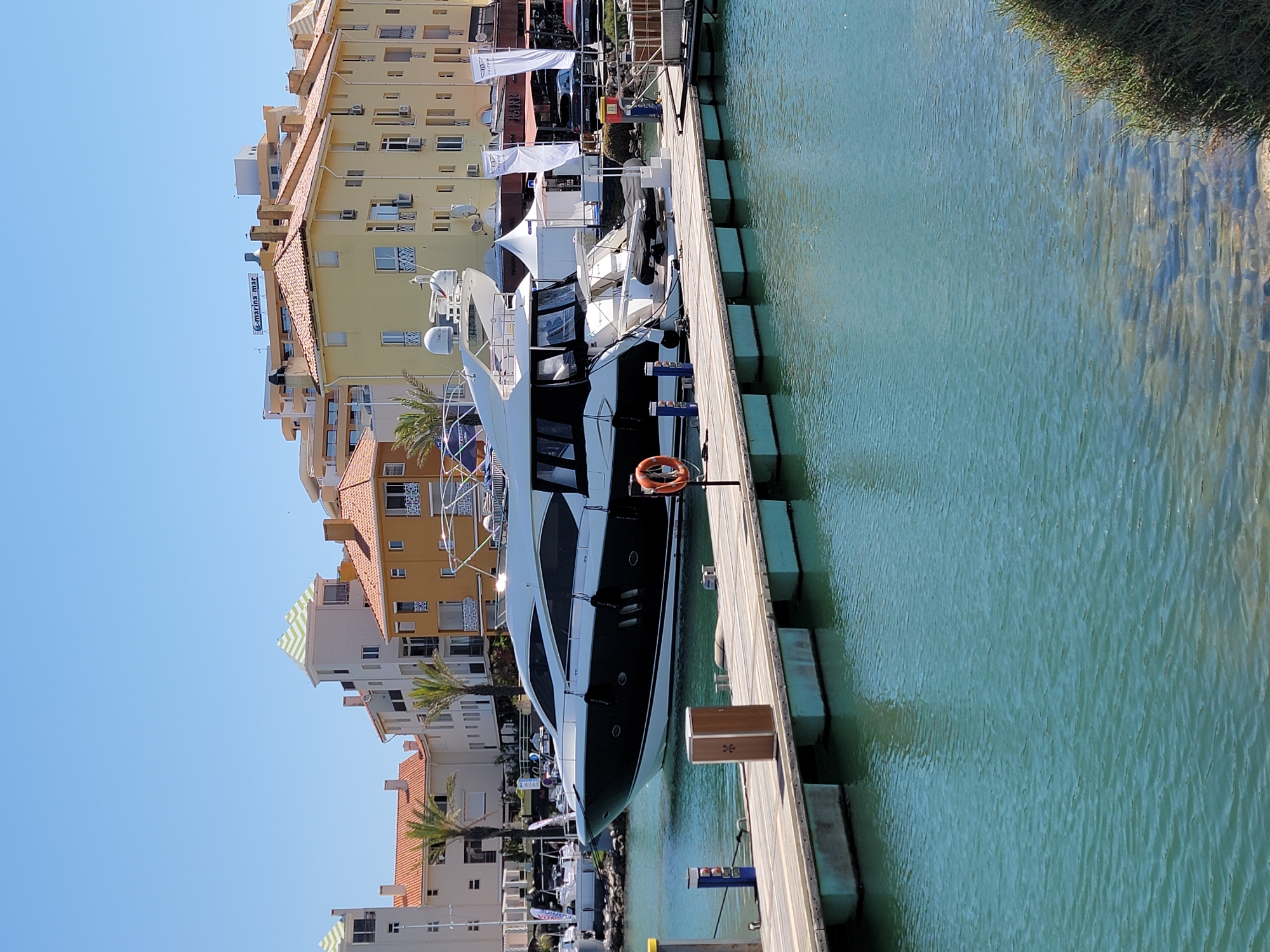 The image depicts a luxury yacht docked at a marina in Quarteira, Portugal, under a clear blue sky. The yacht, predominantly white with a black hull, is moored to a wooden pier. Several buildings with red-tiled roofs and light-colored facades are visible in the background, characteristic of coastal architecture. Palm trees are interspersed among the buildings. The water in the marina is a clear turquoise color. A life preserver is attached to a railing on the pier. The scene conveys a sense of tranquility and affluence associated with a marina setting.