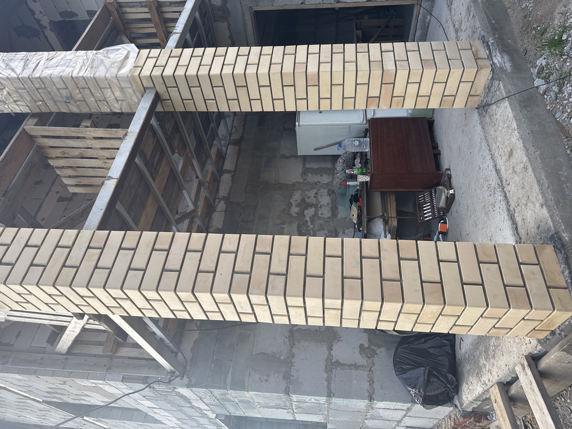 The image shows the construction of brick columns in what appears to be a building under construction. The bricks are light yellow and arranged in a staggered pattern. Some columns are covered in plastic sheeting. The setting looks like a construction site with exposed concrete, wooden supports, and various tools and equipment scattered around. There is a small wooden table and a white refrigerator visible in the background. A black garbage bag sits near the base of one of the columns. The exact location within Perm, Russia cannot be determined from the visual information.