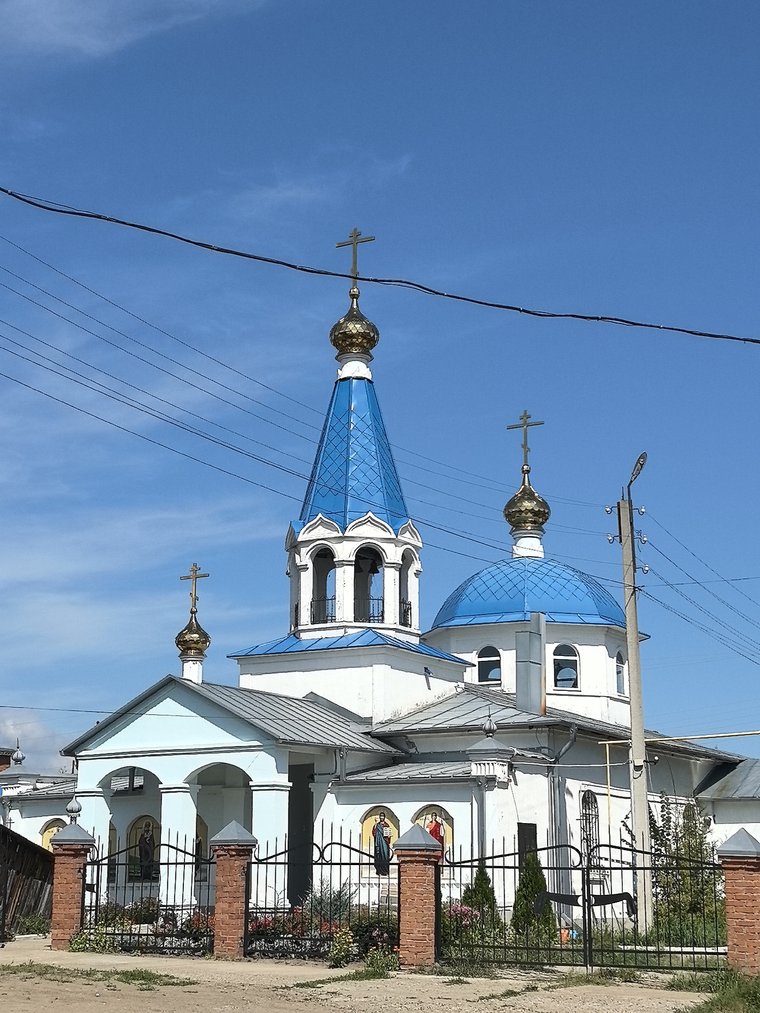 A bright, sunny day illuminates a white and blue domed Russian Orthodox church in Noviy, Russia. The church features multiple blue, metal-roofed domes topped with golden crosses. One prominent dome is a bell tower, and another is a larger, rounded dome. The church is surrounded by a black metal fence with brick pillars. The ground in front of the fence is dirt and gravel with some sparse green vegetation. Behind the church, power lines crisscross the clear blue sky, and a utility pole with a street light is visible to the right of the church. The overall impression is of a peaceful, rural religious building.