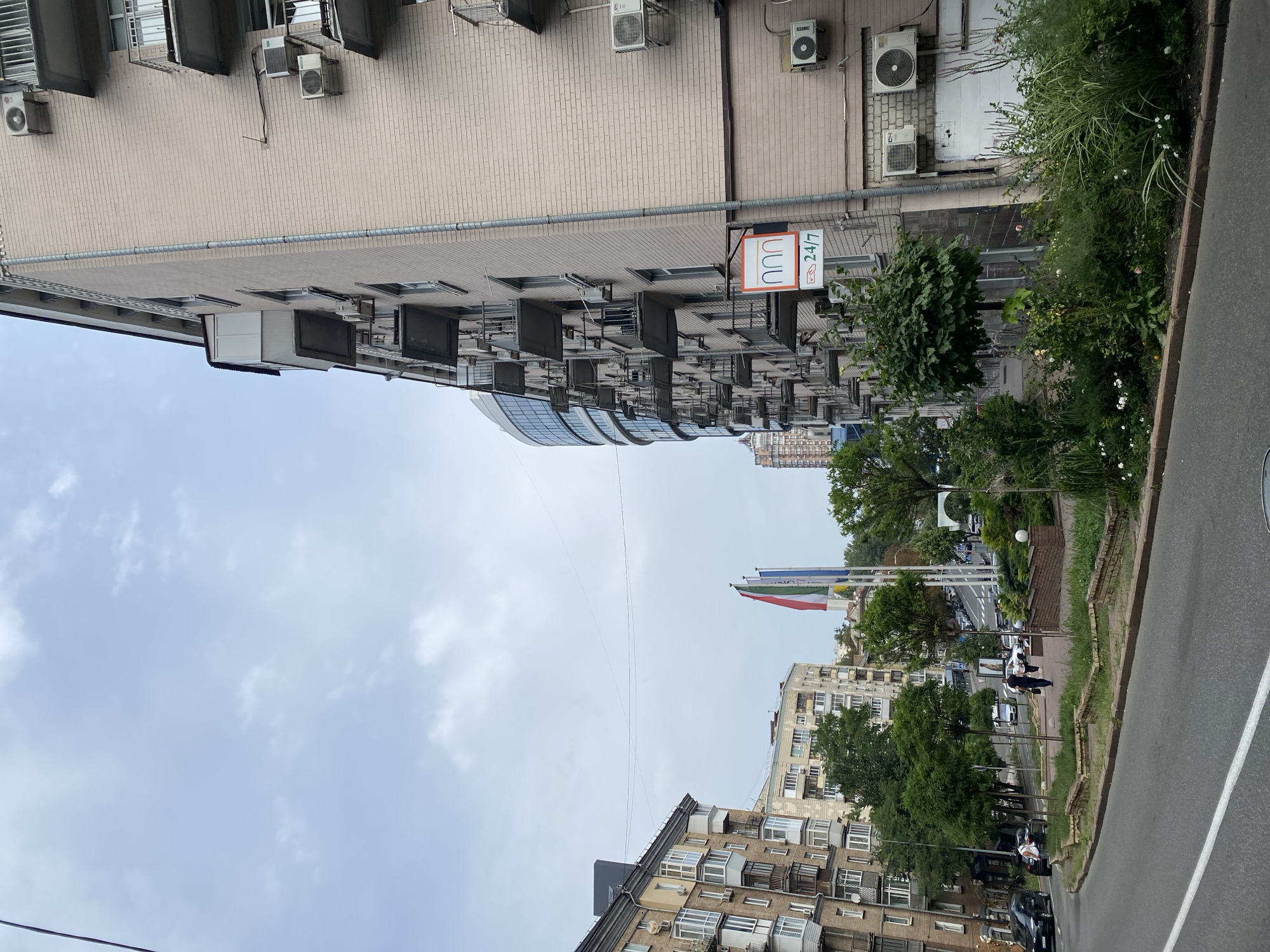 A street view in an urban environment is depicted on an overcast day. The foreground features a grey asphalt road with a white line marking. To the right, a raised planter bed with overgrown green plants and flowers lines the sidewalk. Adjacent to the planter is a pale brick building with multiple balconies and several air conditioning units mounted on its exterior. A sign with the text "n n n 24/7" is visible on the building's facade.
Further back, a multi-story apartment building with many windows and balconies stands on the left side of the street. A street lined with trees runs parallel to the road, with cars parked and driving along it. In the mid-ground, several flags, including red, white, and blue, are displayed on flagpoles. Another building with a curved glass facade is visible in the distance. The sky is cloudy and pale blue.