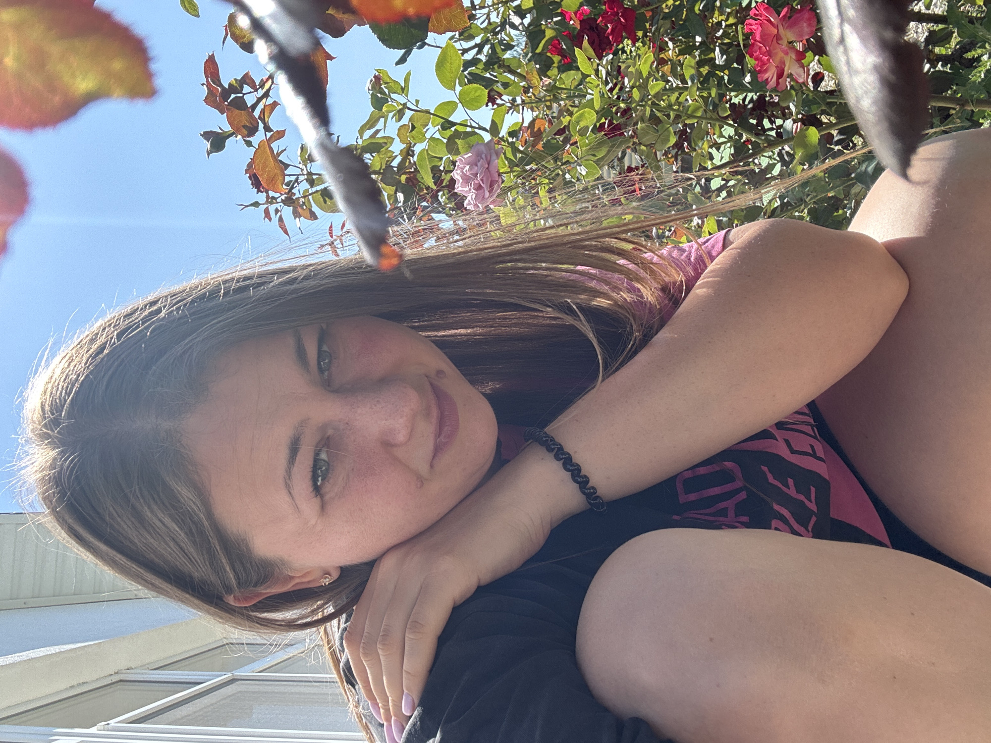The image shows a close-up of a woman outdoors on a sunny day. She has long brown hair and is looking at the camera with a slight smile. She is wearing a black shirt with pink lettering that reads "BAD GIRL". Her left arm is bent and resting on her shoulder, and her right arm is bent and resting on her leg. In the background, there are rose bushes with pink and red flowers. The scene appears to be in a residential area, with a house visible on the left side of the frame. The woman's skin appears slightly tanned and she has freckles across her nose and cheeks. The lighting suggests it is daytime and the sun is shining brightly.