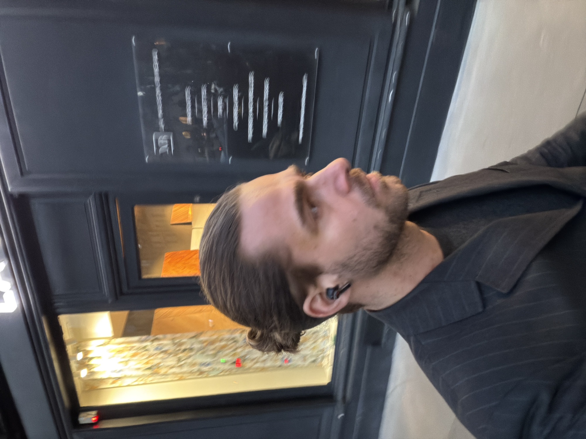 The image depicts a male subject walking in profile on a city sidewalk next to a dark-colored building facade, characteristic of a commercial street in Paris, France.
The subject, positioned in the foreground and facing right, has brown hair styled in a bun at the back and features a beard and mustache. He wears a dark pinstripe suit jacket over a dark undershirt. A black wireless earbud is visible in his left ear. His gaze is directed forward and slightly downward.
The background comprises a building with a dark facade. To the subject's left, a brightly lit display window showcases a patterned fabric or material, featuring a light background adorned with abstract yellow, orange, and blue designs. Small red and green indicator lights are visible within this display. Partial letters "LA" are discernible from a sign positioned above this window at the top of the building. To the subject's right, a non-transparent section of the facade, possibly a door, features a reflective plaque. The letters "NR" are visible on the upper left of this plaque, with several lines of blurred, illegible text below, likely indicating information such as hours of operation or services. The pavement beneath the subject is a light grey. The scene suggests an upscale urban setting.