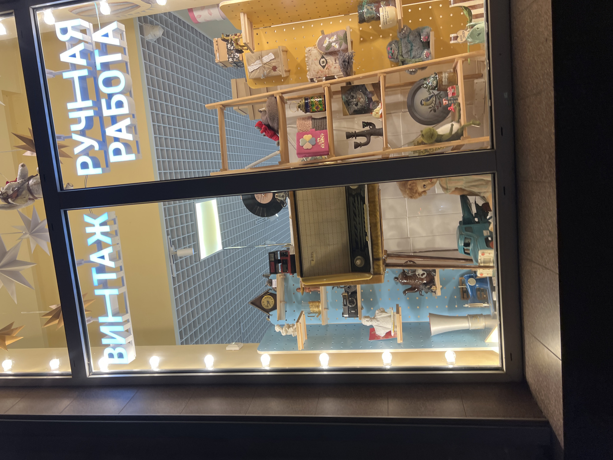 This is a nighttime, eye-level, outdoor shot of a storefront window display. The window is illuminated from within, showcasing various vintage and handmade items. The sign above the display reads "ВИНТАЖ РУЧНАЯ РАБОТА", which translates to "Vintage Handmade". The display features shelves filled with objects like old radios, cameras, dolls, decorative plates, figurines, and gift-wrapped packages. A wooden ladder is positioned to the right, also holding various items. The window itself is framed by dark gray exterior walls and has a tiled windowsill in the foreground. The scene is likely located in the city of Krasnodar, Russia, given the Cyrillic signage.