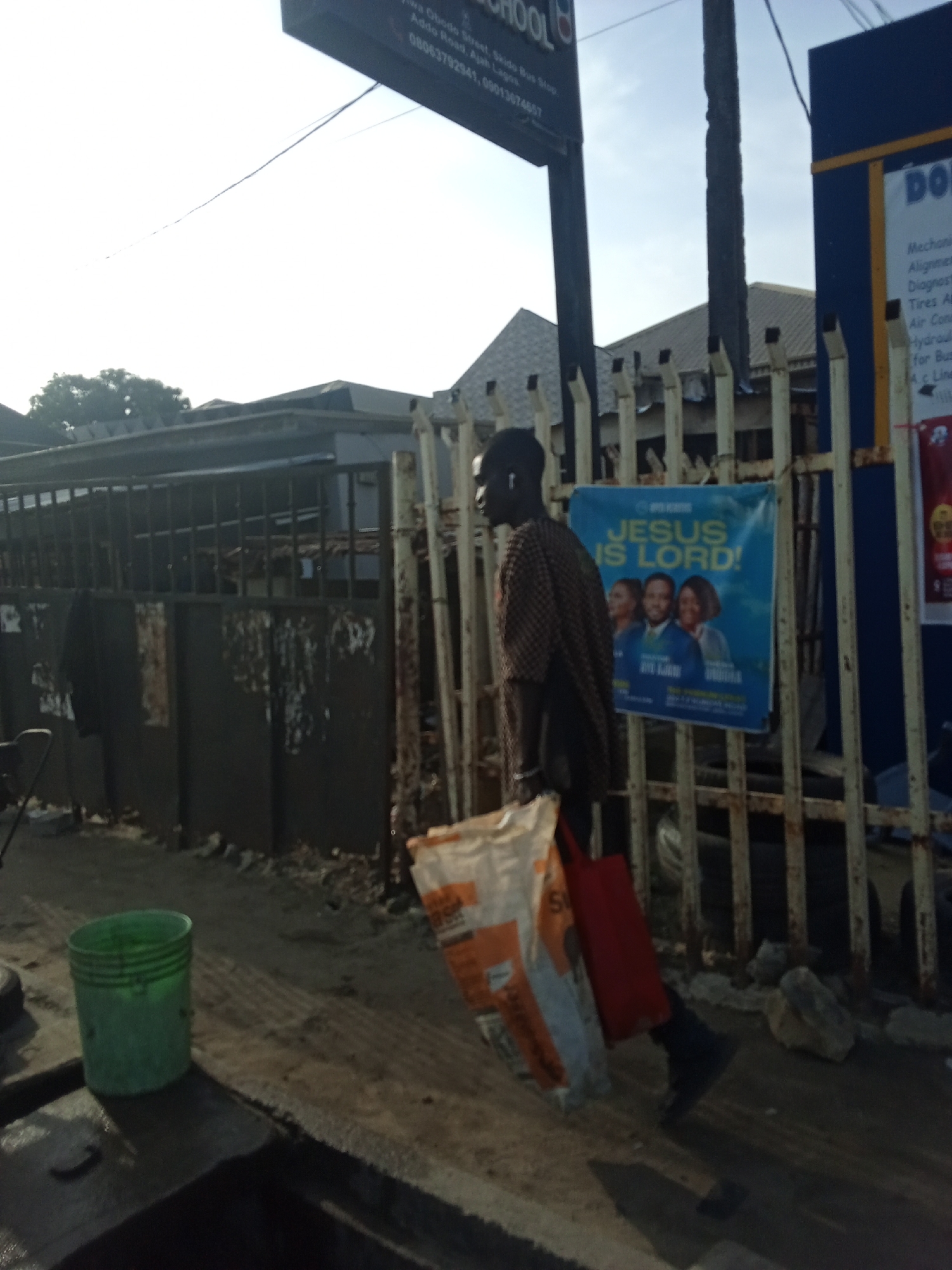 A man walks down the street in Ado, Nigeria, carrying a large white and orange bag in one hand and a red bag in the other. He passes a metal fence with a "Jesus is Lord" poster attached. The fence also has a sign for auto repair services and a school. A green bucket sits on the ground near a black, oily surface. The location is identified by a sign stating "Awa Ohodo Street, Skido Bus Stop, Addo Road, Ajah Lagos," along with phone numbers. The scene appears to be a typical street view in the area.