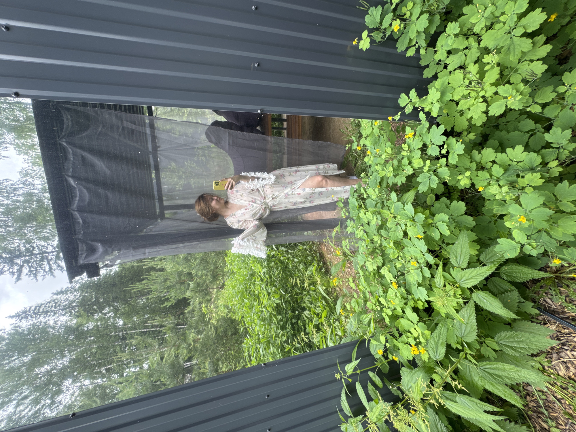 A woman stands in a gazebo with sheer curtains, taking a mirror selfie with her phone. She is wearing a floral robe and has her hair up. The gazebo is surrounded by lush greenery and trees, suggesting a natural, outdoor setting. The image does not provide any information that would allow for its location to be identified as Bakhtiyary, Russia.