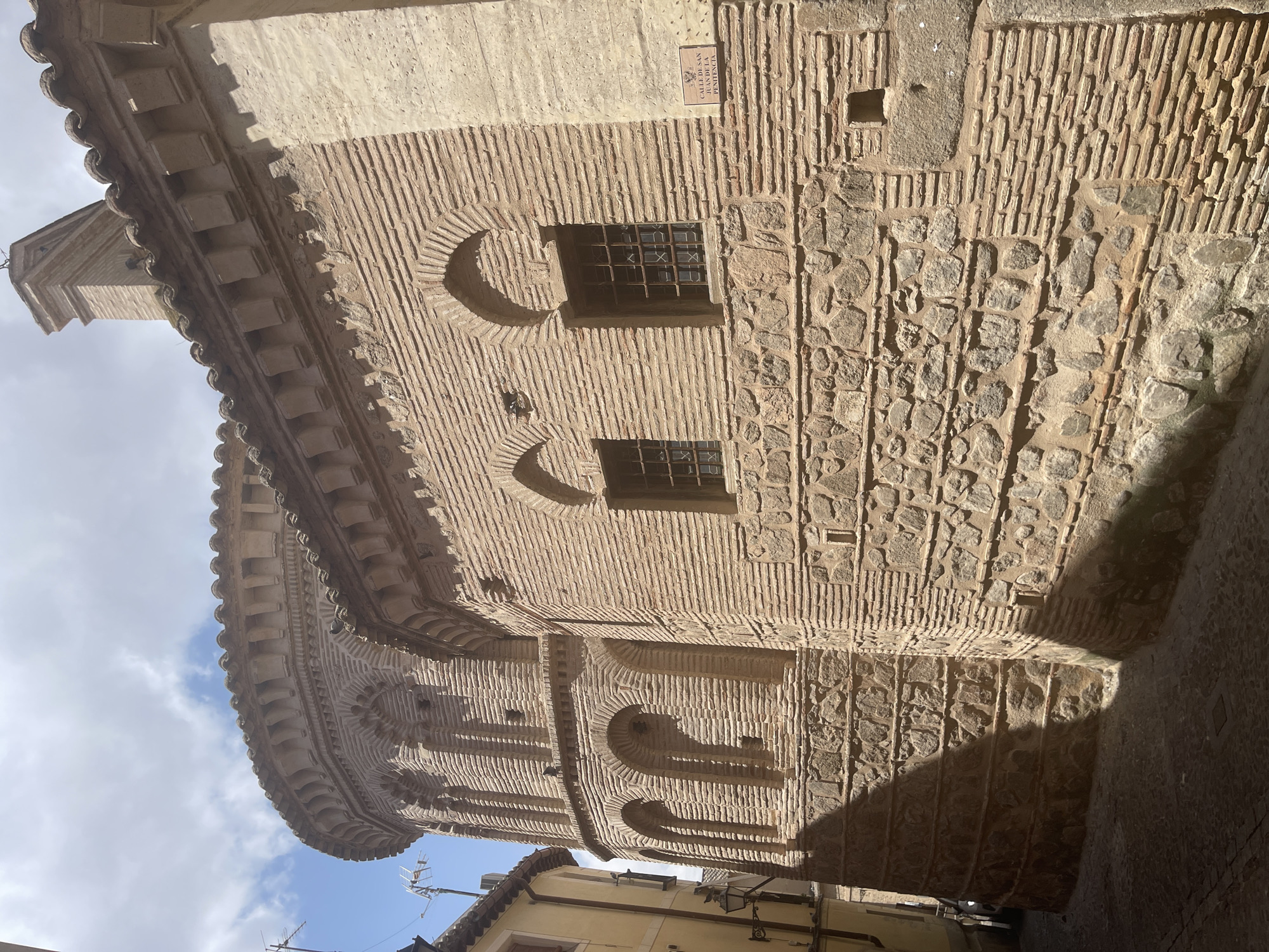 The image shows a close-up, eye-level view of the exterior of a historic building made of brick and stone, located in Toledo, Spain. The building features rounded architectural elements, arched windows with metal grates, and a decorative frieze along the roofline. A street sign is visible on the wall, reading "CALLE DE SAN JUAN DE LA PENITENCIA". The building is situated on a narrow cobblestone street, with shadows cast by the sun indicating it is daytime. The sky above is partly cloudy. No people or actions are depicted in the scene.