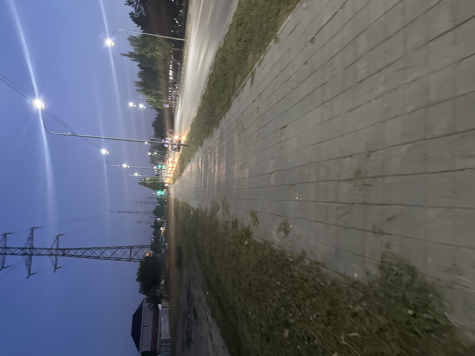 The image depicts a wide paved pedestrian walkway running alongside a road at dusk in the city of Kostanay, Kazakhstan. A row of streetlights illuminates the walkway, casting long shadows and creating light streaks across the sky. The road, which appears to be a multi-lane highway, has several cars driving on it, their headlights and taillights visible. On the left side of the walkway, there is a strip of dry grass and dirt, with a distant house and a large power line tower visible in the background. The sky is a deep blue, transitioning into lighter hues towards the horizon, indicating early evening. The overall scene suggests an urban or semi-urban environment.